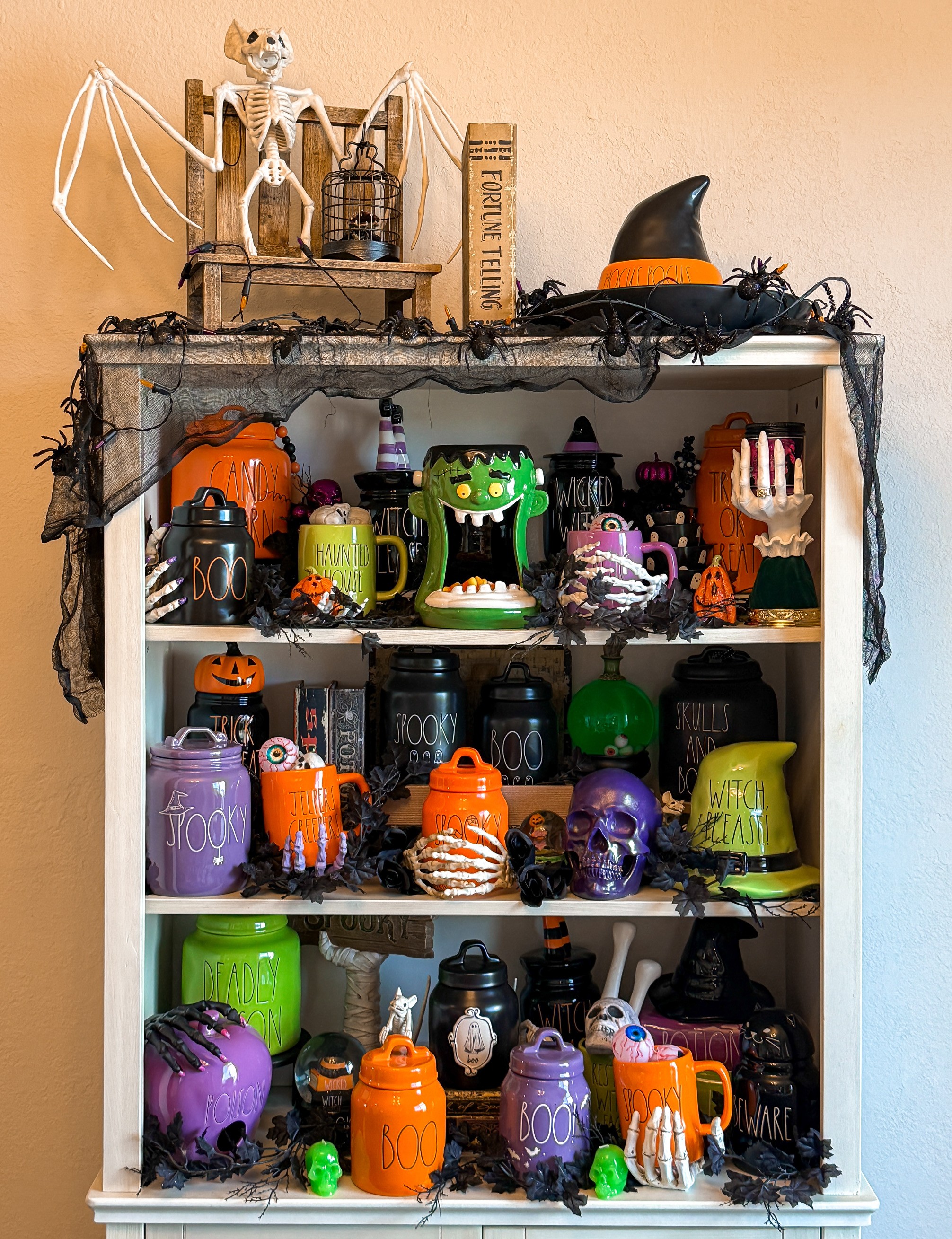 Decorated shelf with Halloween-themed items, including pumpkins, a witch hat, skulls, and spooky decorations, against a beige wall.