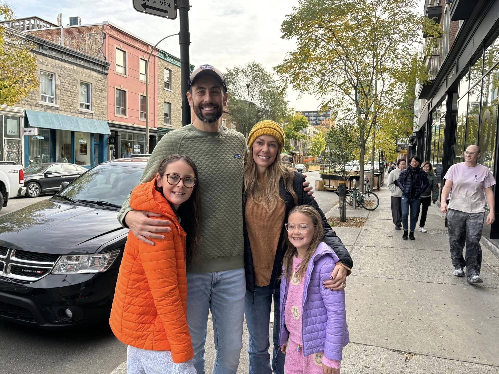 Group of four people smiling on a city sidewalk, with buildings and trees in the background.
