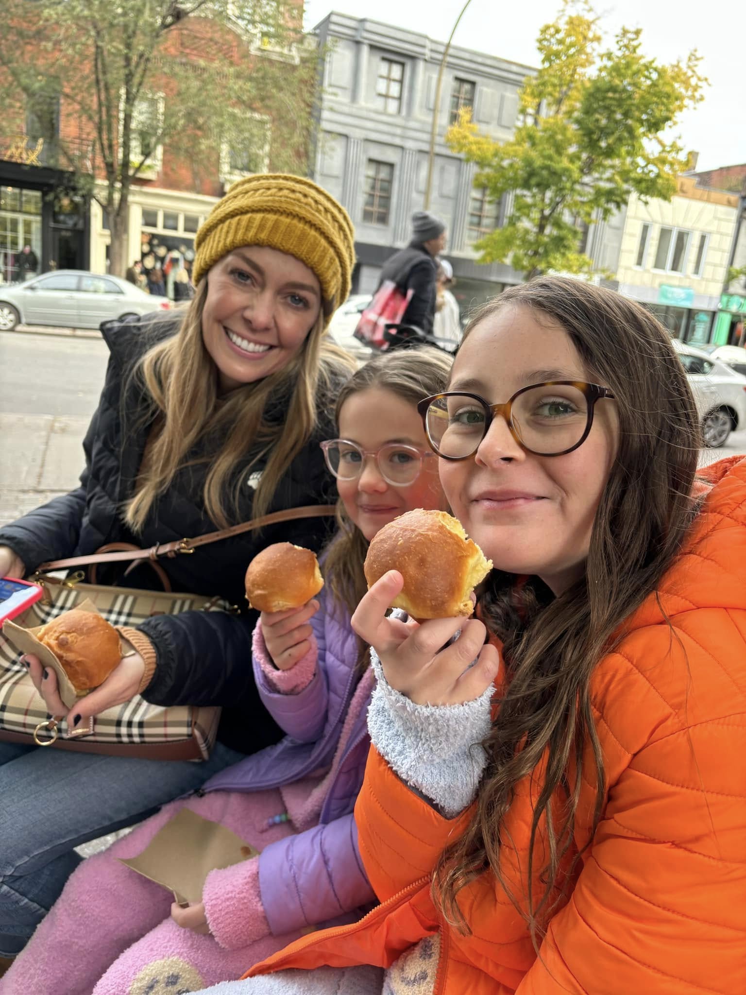 Three women sitting outdoors, smiling, holding donuts, with trees and buildings in the background.
