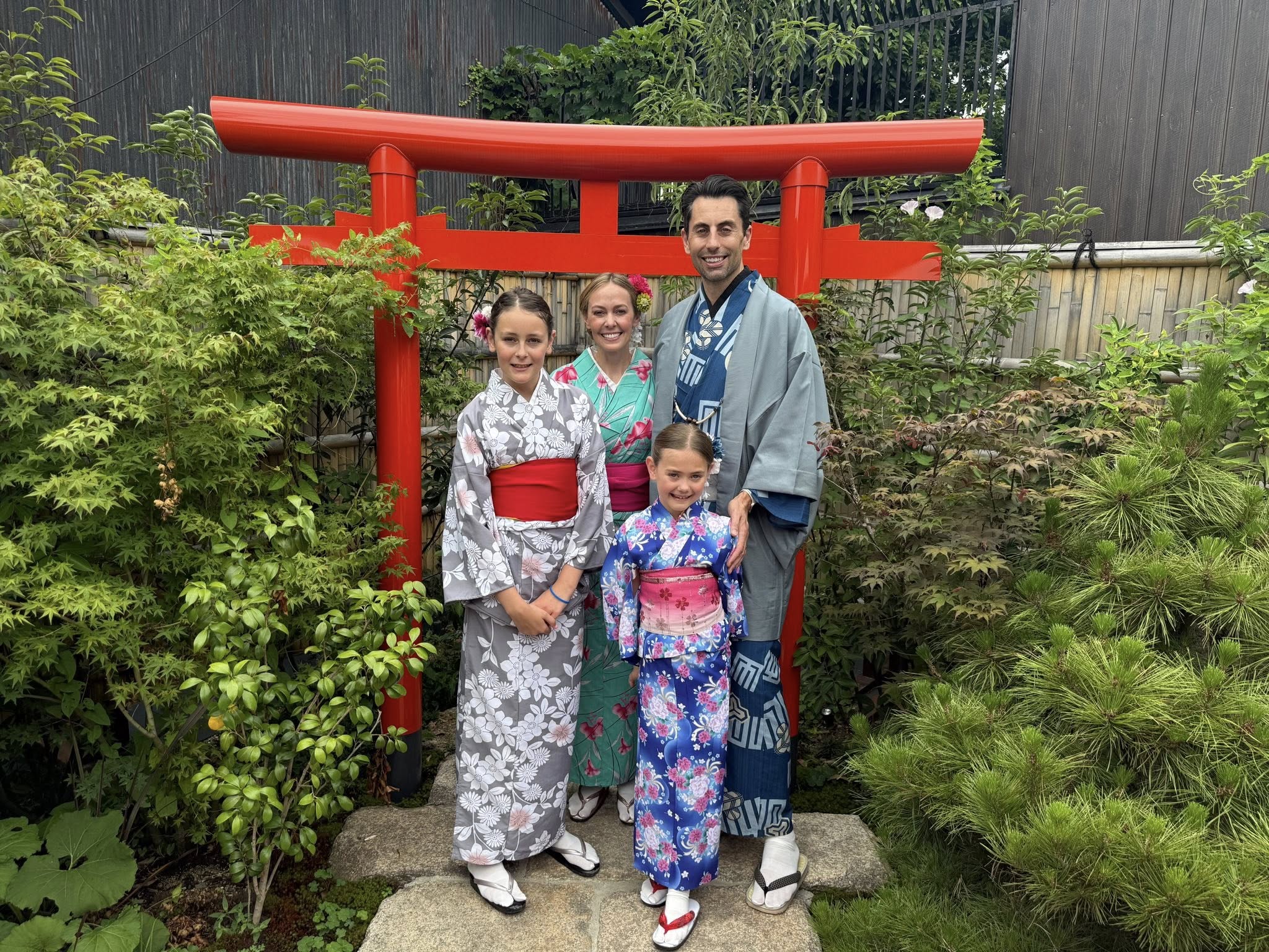 Family of four in traditional Japanese clothing standing in front of a red torii gate, surrounded by greenery.