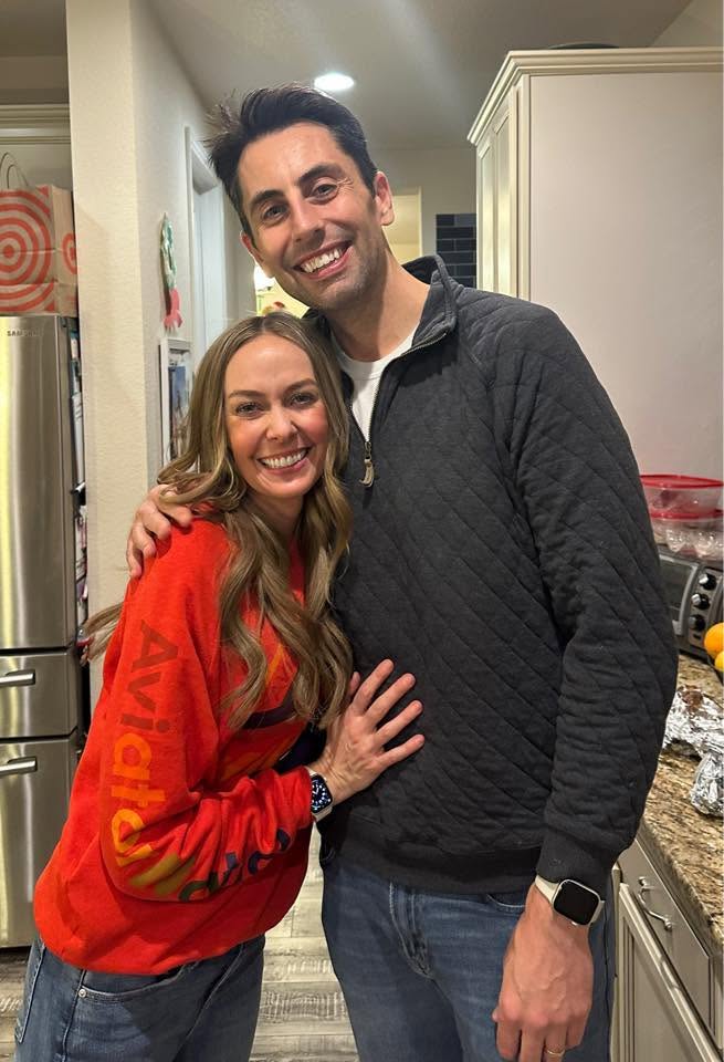 Two people smiling, one with long hair in a red sweatshirt, the other with short dark hair in a dark jacket, standing in a kitchen.