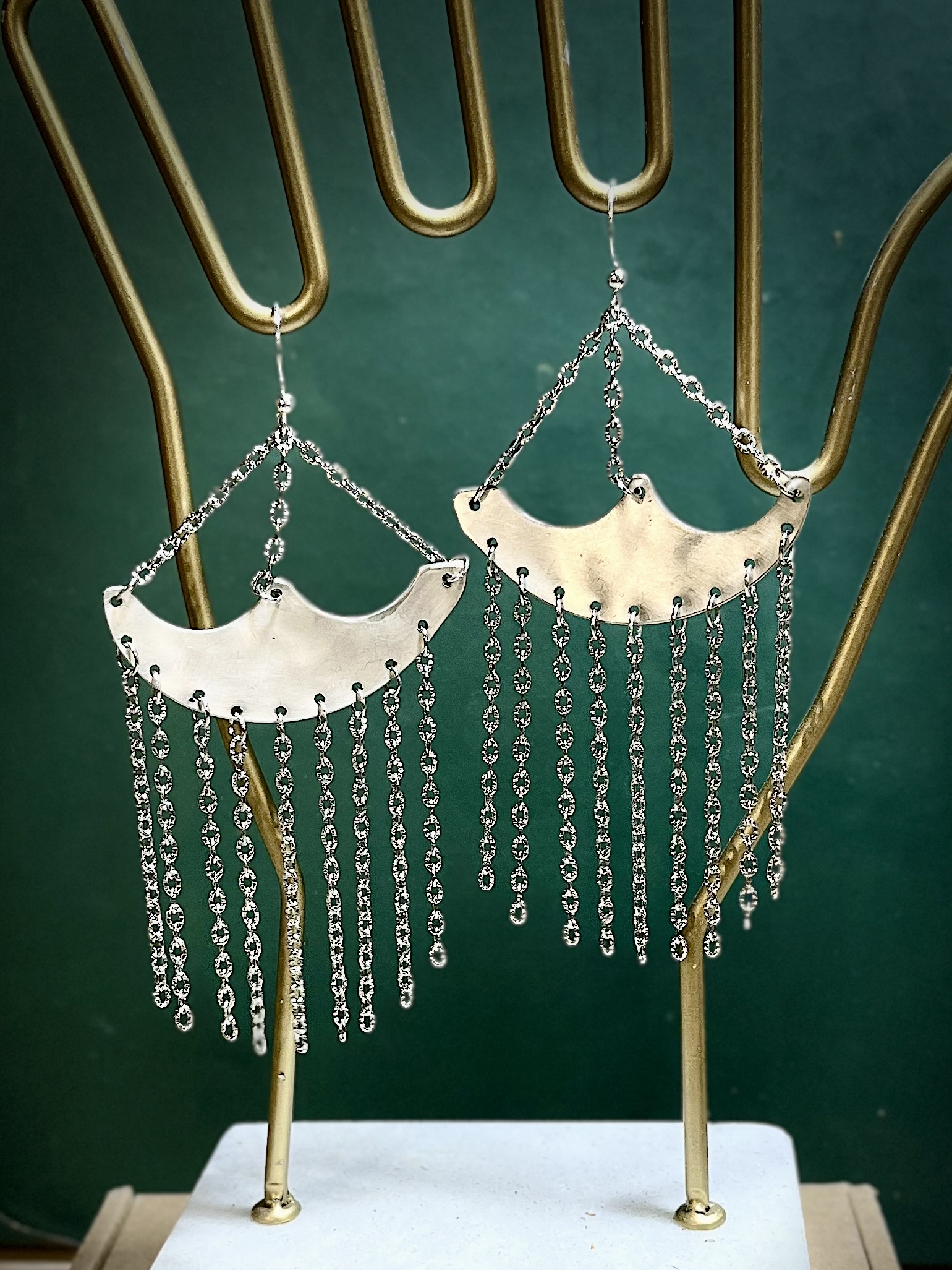 Pair of silver earrings with chain fringe hanging from curved, crescent-shaped bases, displayed on a gold stand against a green background.
