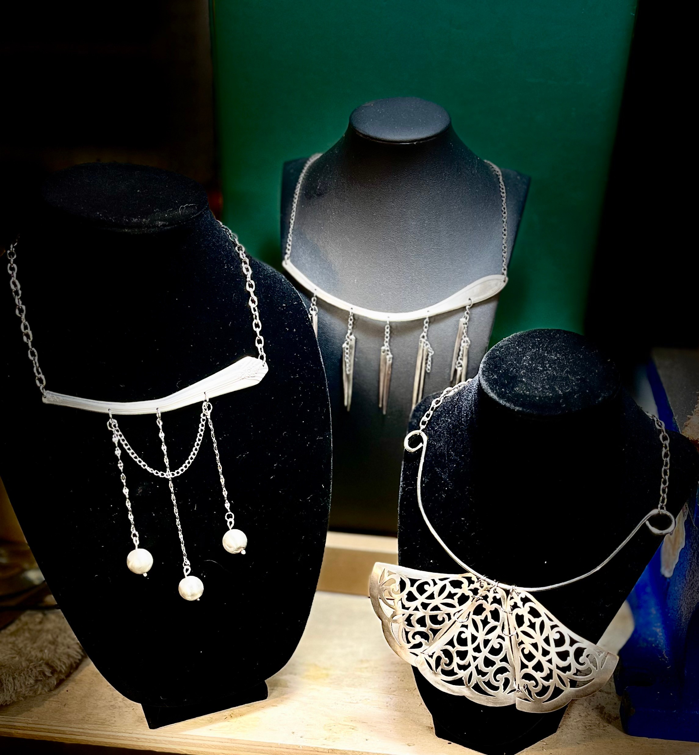 Three necklaces displayed on black and gray busts, with intricate designs and hanging pendants, on a wooden surface.