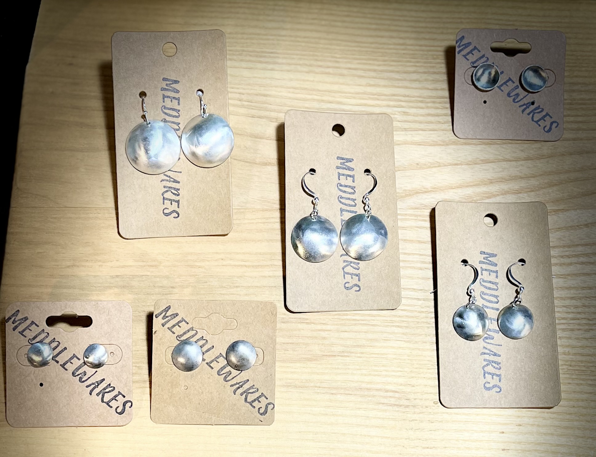 Six pairs of earrings on beige tags, displayed on a light wooden surface.