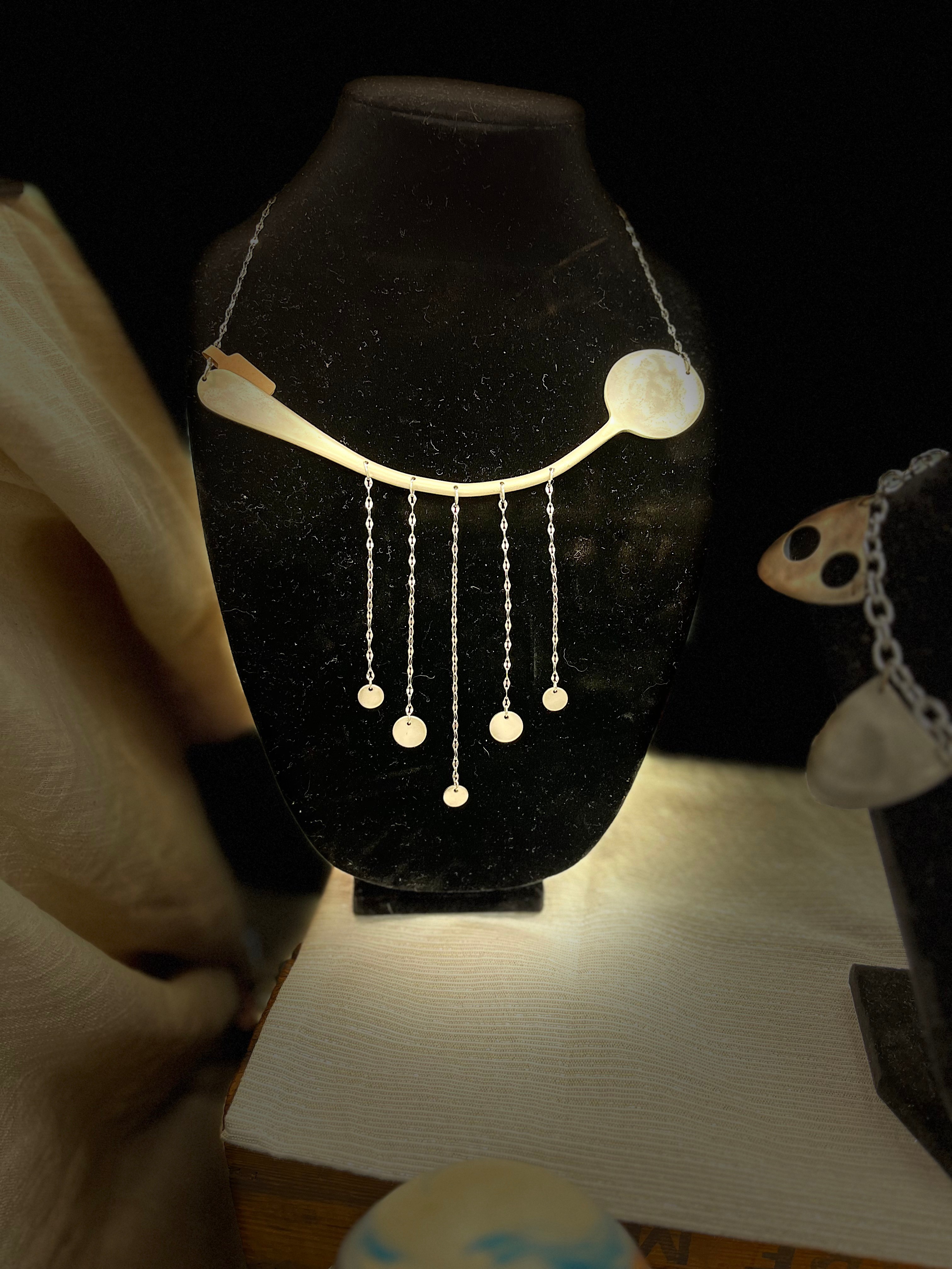 Elegant necklace with circular and linear pendant elements displayed on a black stand, illuminated against a dark background.