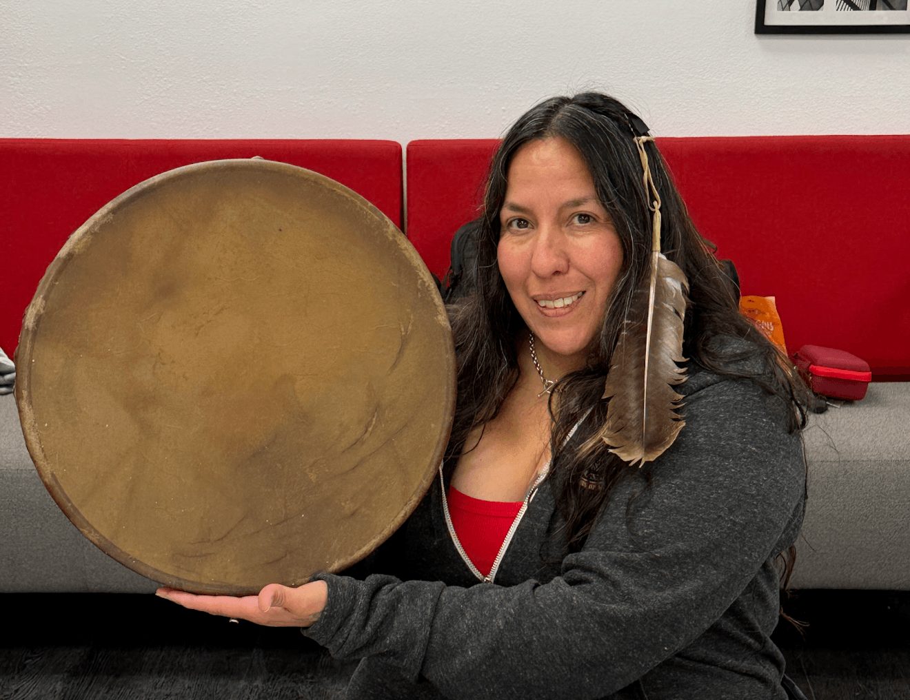 Woman smiling holding a large round drum with a feather in her hair, sitting in a room with red and gray background.