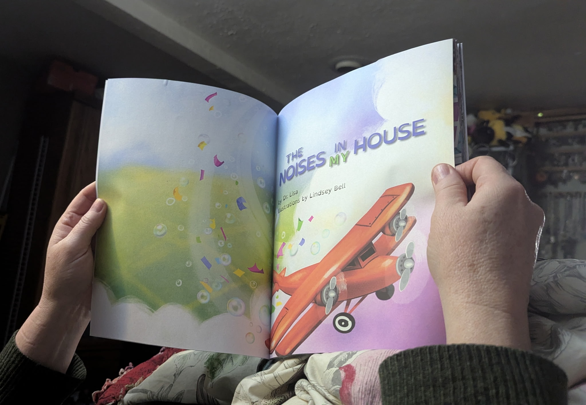 Open children's book with colorful illustrations and text, held by two hands, in a room with a ceiling and furniture.