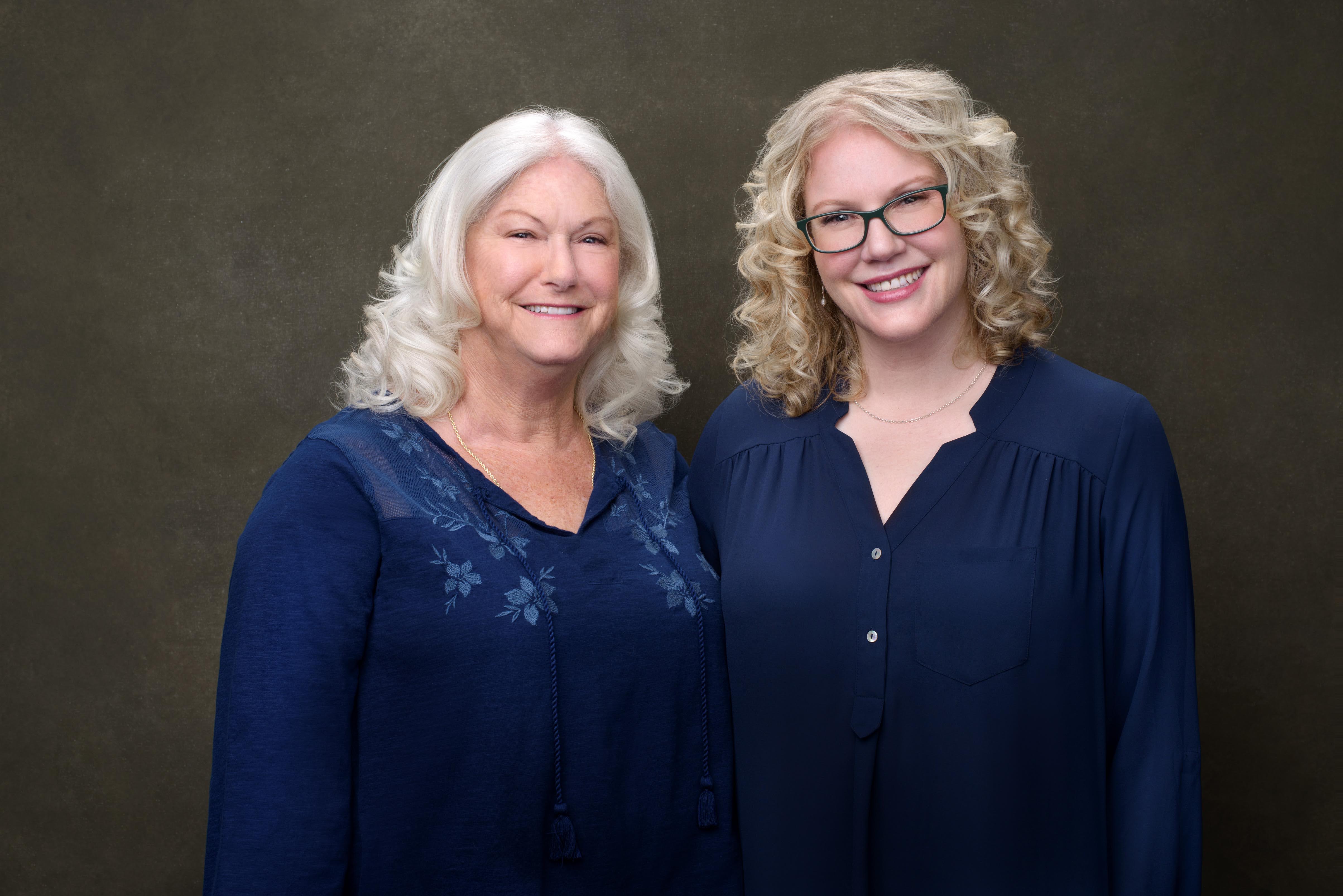 Two women with light hair smiling, wearing blue tops, standing against a dark background.