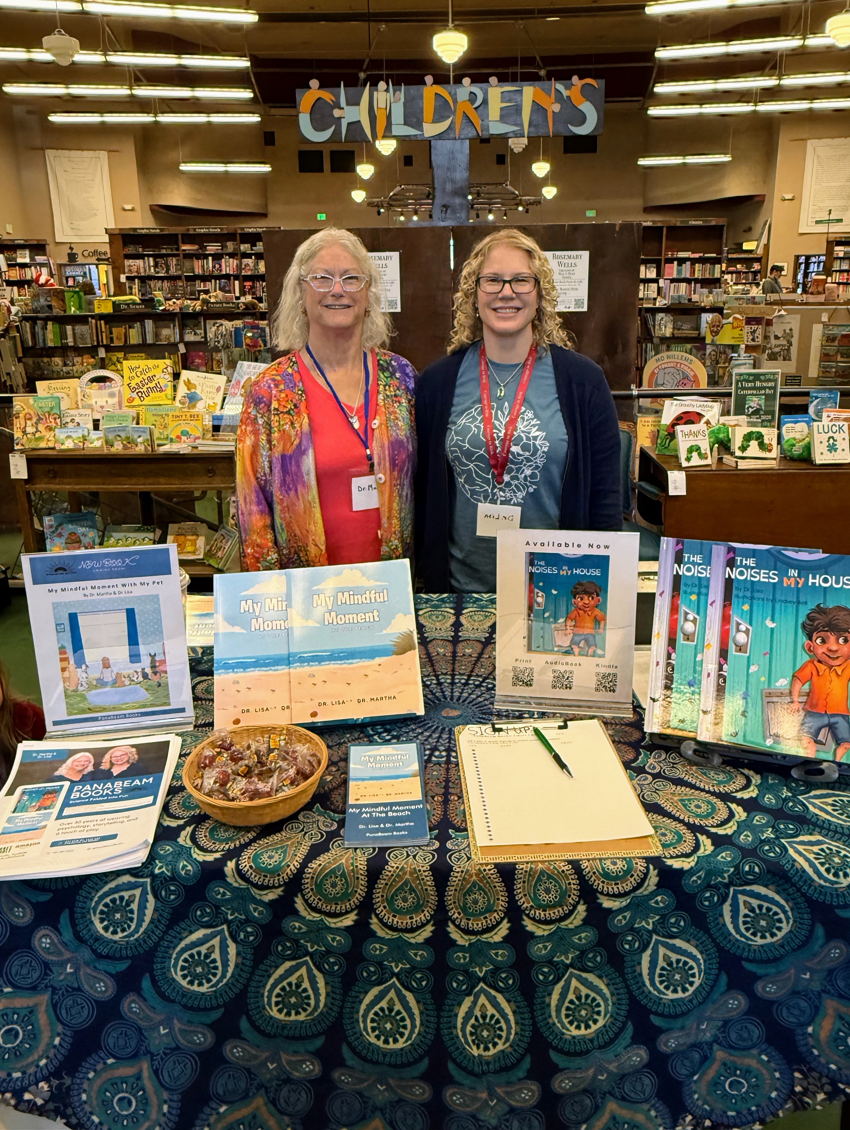 Two women stand behind a table with books and a bowl, in a library with a children's section sign overhead.