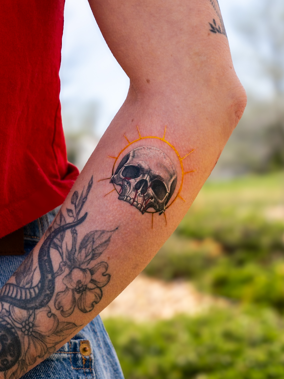 Person's arm with a skull tattoo surrounded by an orange sunburst, and a snake tattoo on the forearm, outdoors.