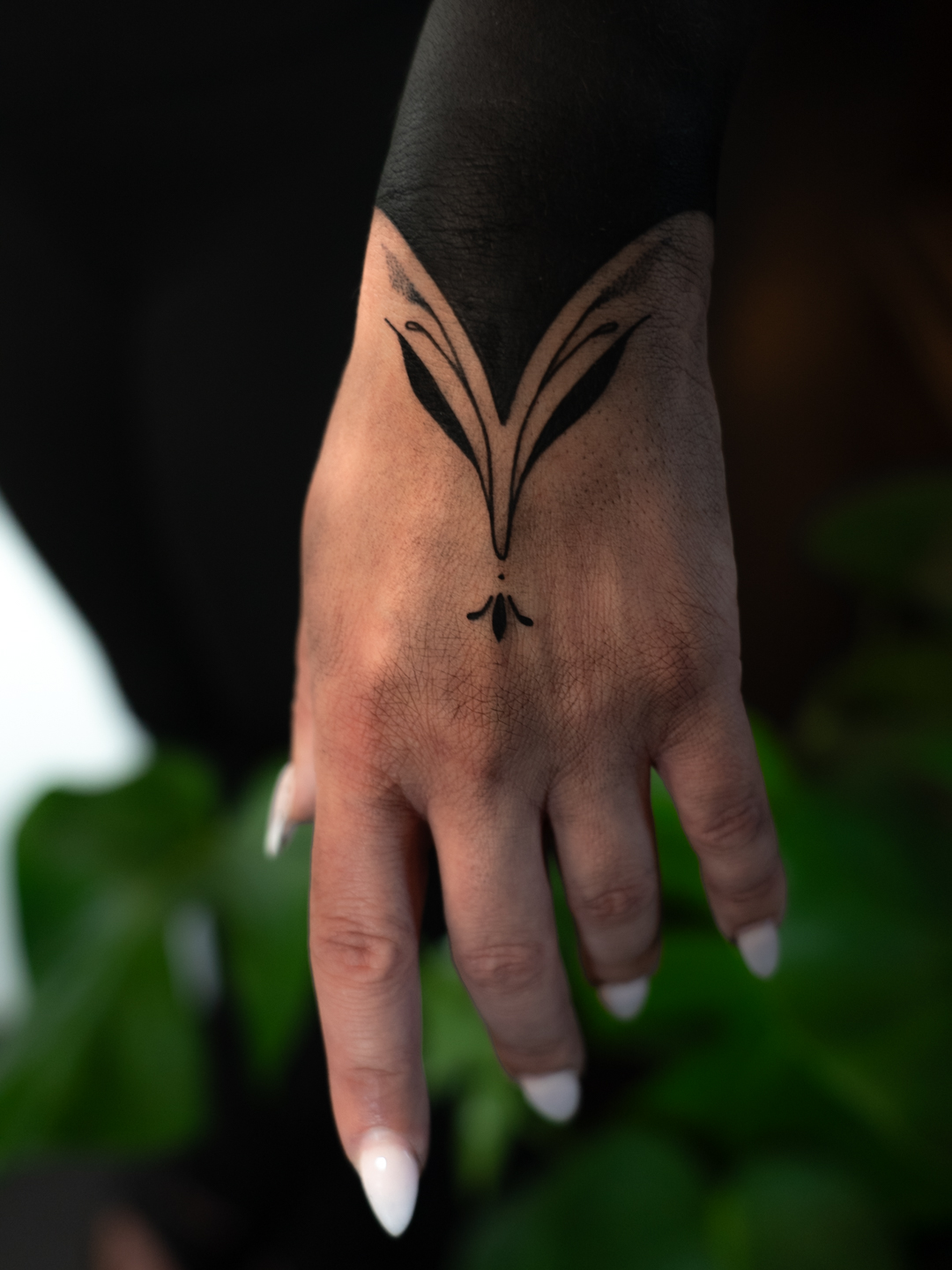 Hand with a butterfly tattoo on the back, black sleeve, and white nail polish, against a dark background with green leaves.