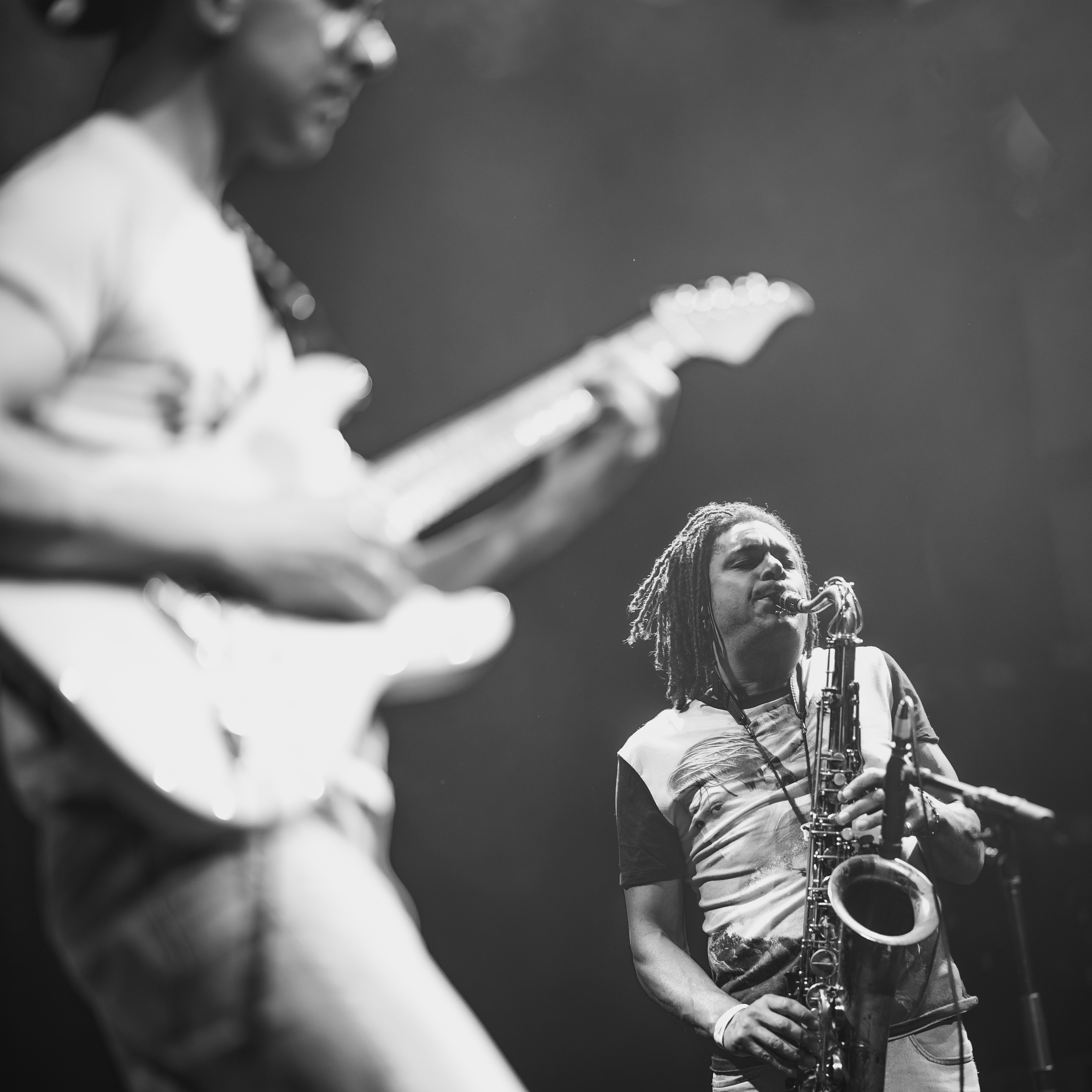 Two musicians perform on stage, one playing guitar and the other playing a saxophone, with a dark background.