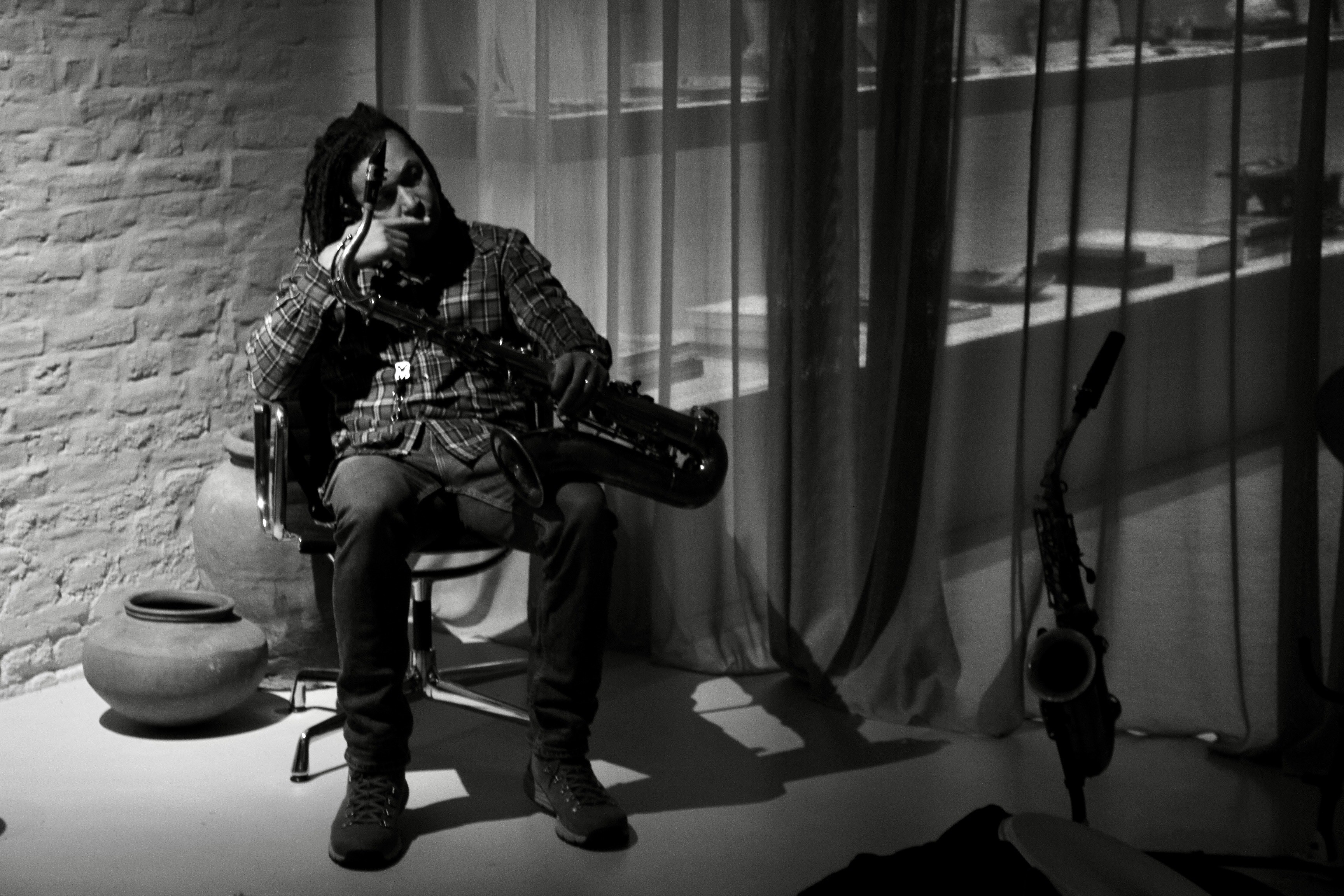 Person sitting on a chair, holding a guitar, with a curtain and shelves in the background, in black and white.