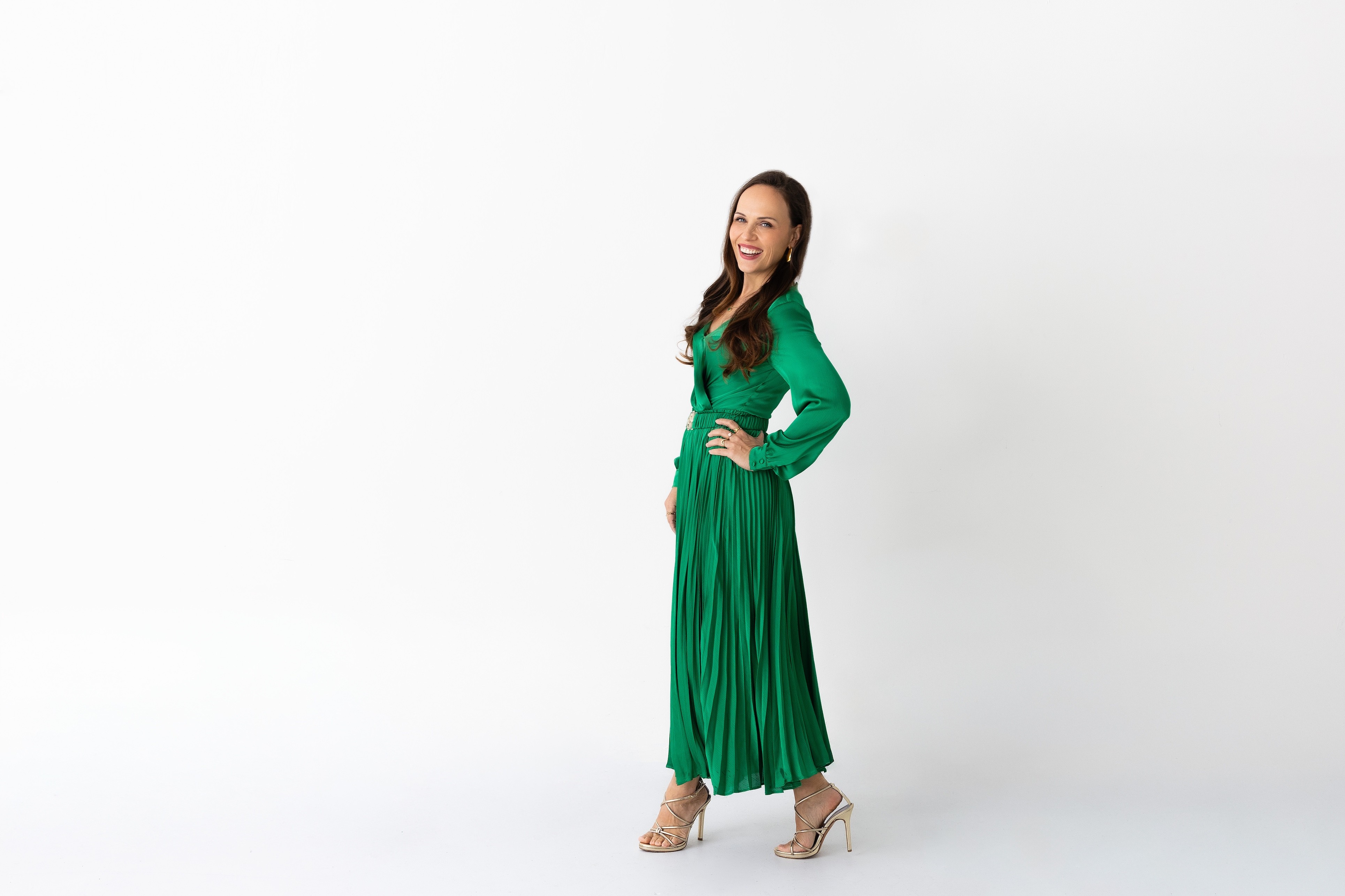 Woman in green dress standing with hand on hip, smiling, against a plain white background.