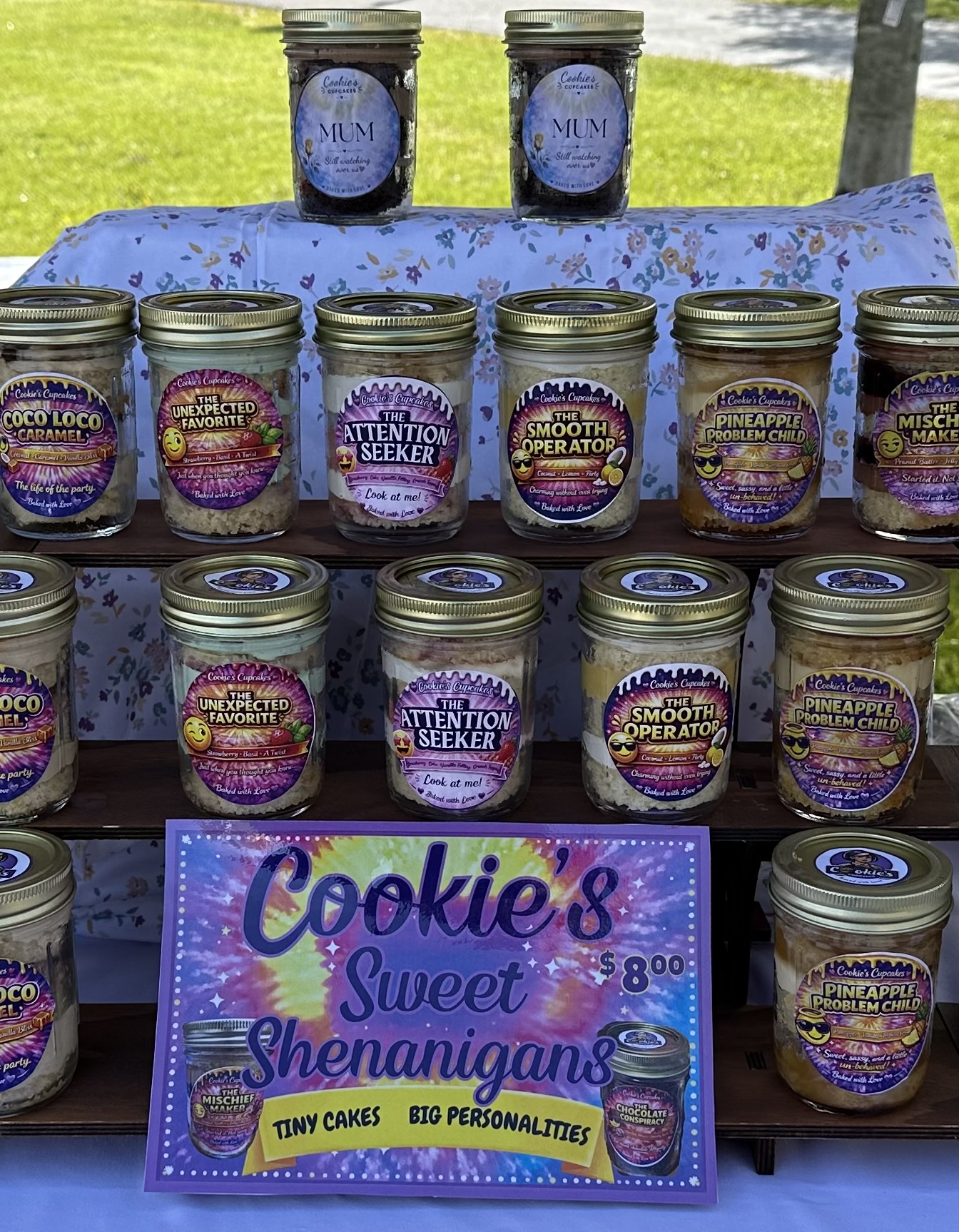 Display of small jars of cookie and sweet-themed products on a table outdoors, with a sign reading 