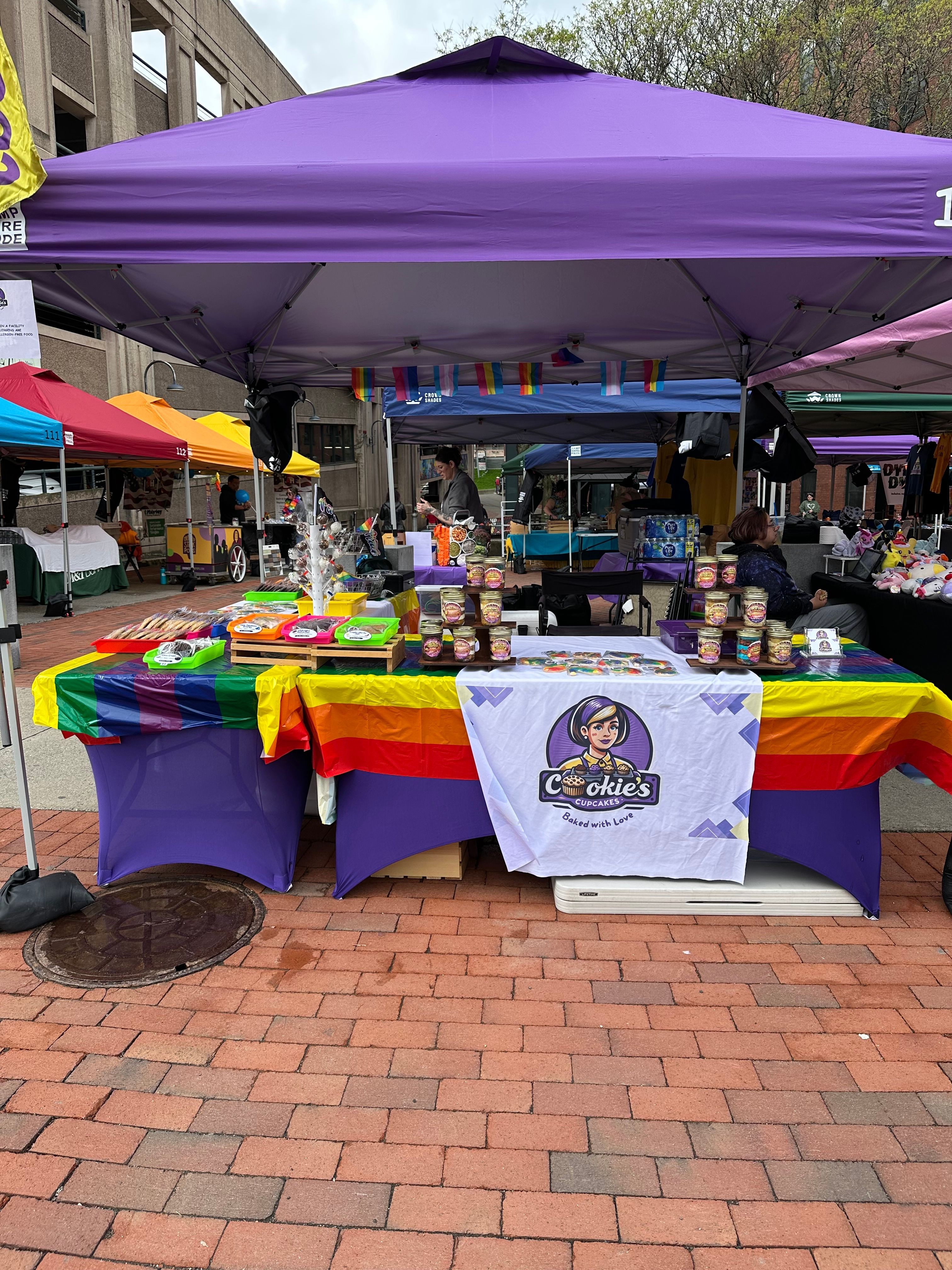 Table with rainbow flag and LGBTQ+ symbols, under a purple canopy, on brick pavement, with people in background.
