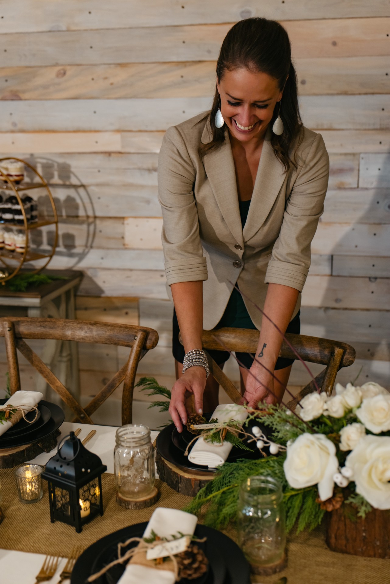 Woman arranging a table with flowers and decorations, smiling, in front of a wooden wall, with chairs and decorative items nearby.