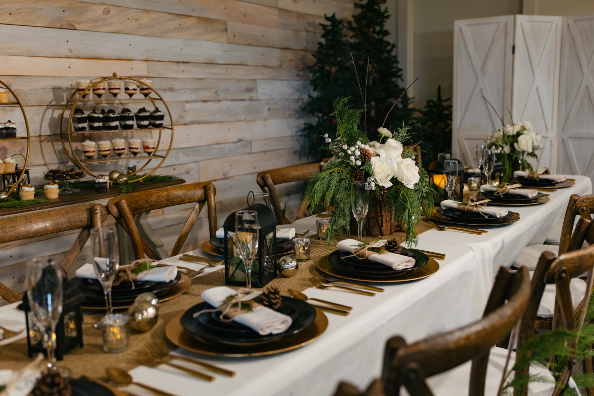 Long dining table set with black plates, gold utensils, white napkins, and floral centerpieces, in a rustic decorated room.