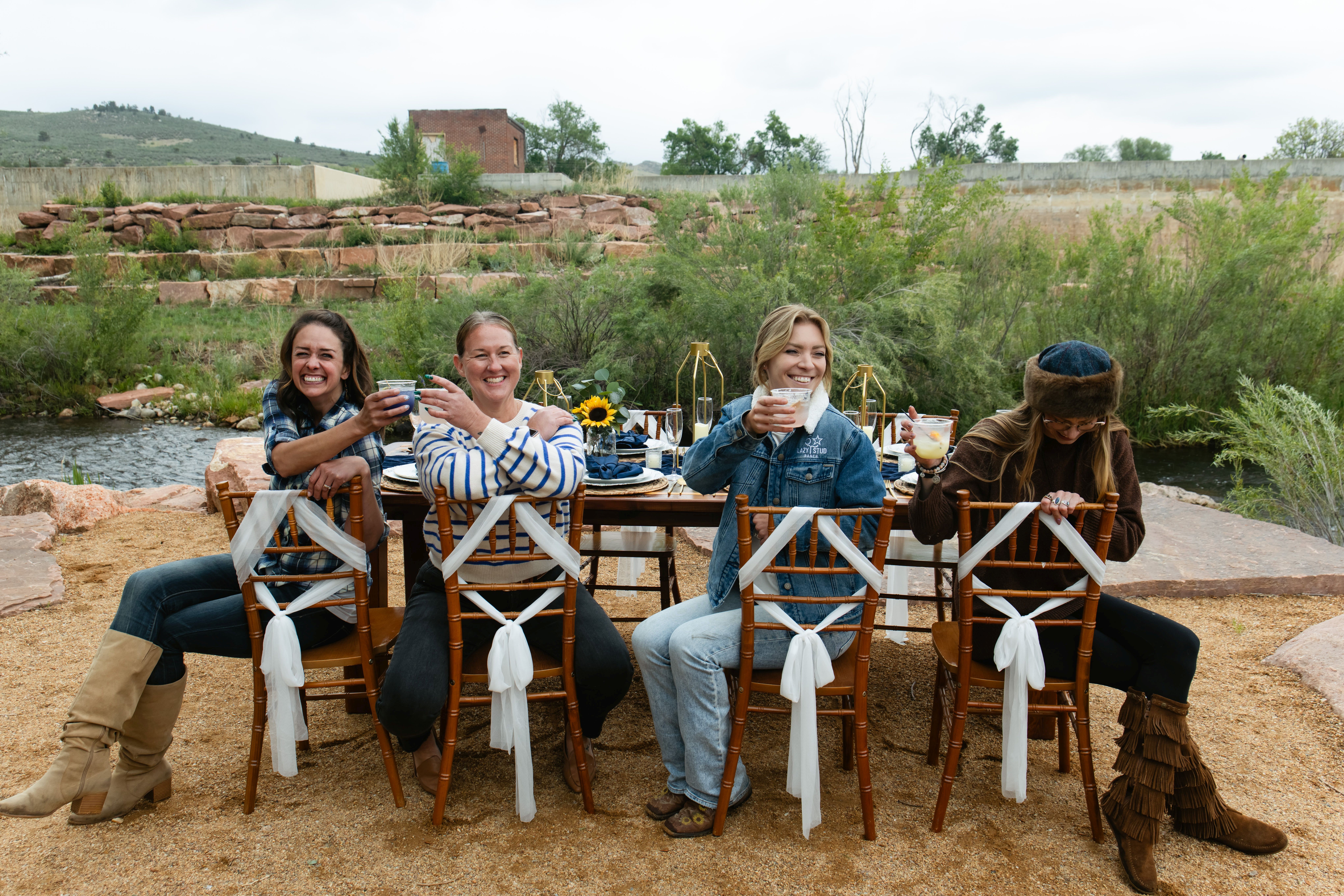 Four women sit at a table outdoors with a river and greenery in the background, smiling and enjoying drinks.