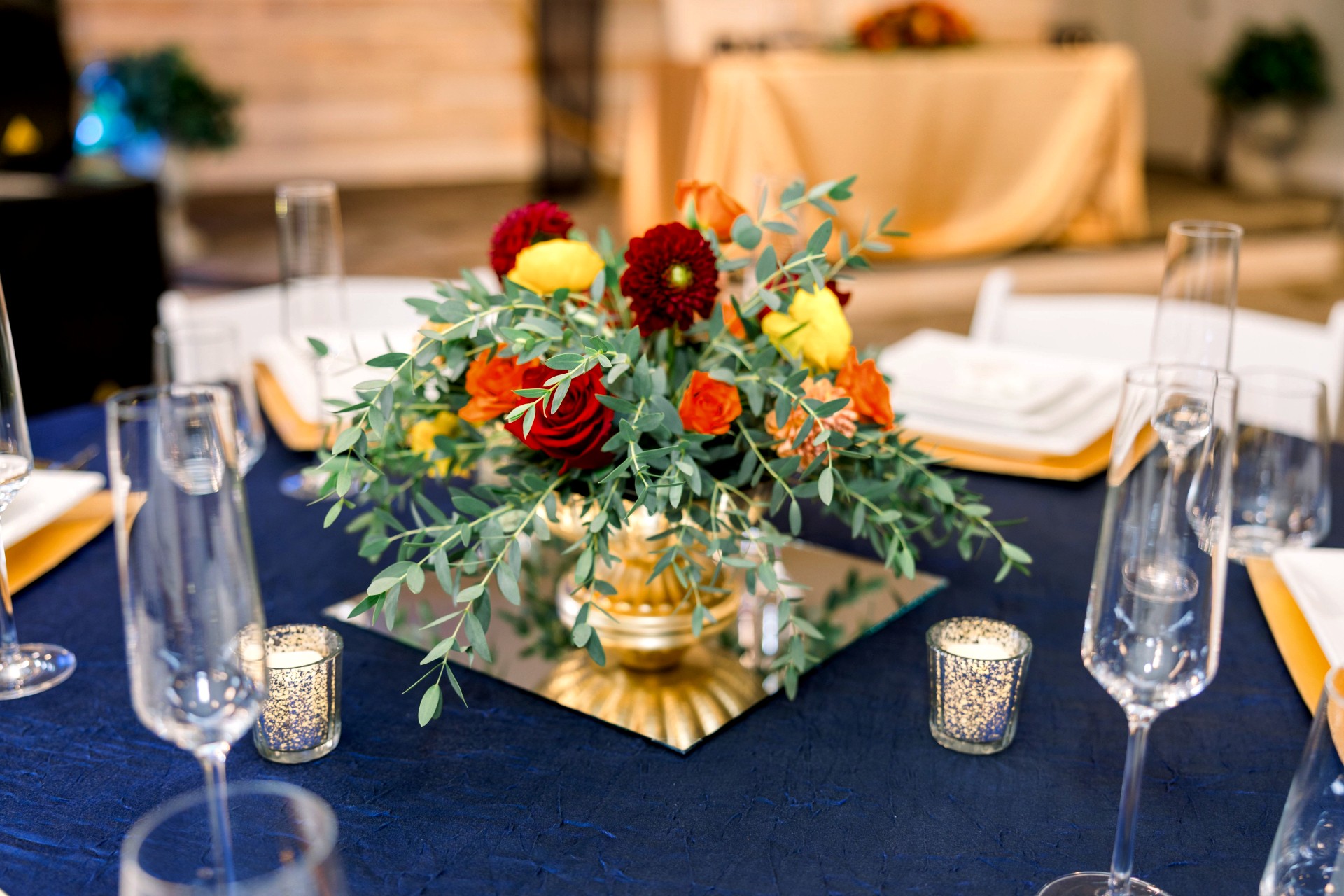 Elegant table setting with a floral centerpiece, glasses, and candles on a dark tablecloth, in a well-lit room with a sofa in the background.