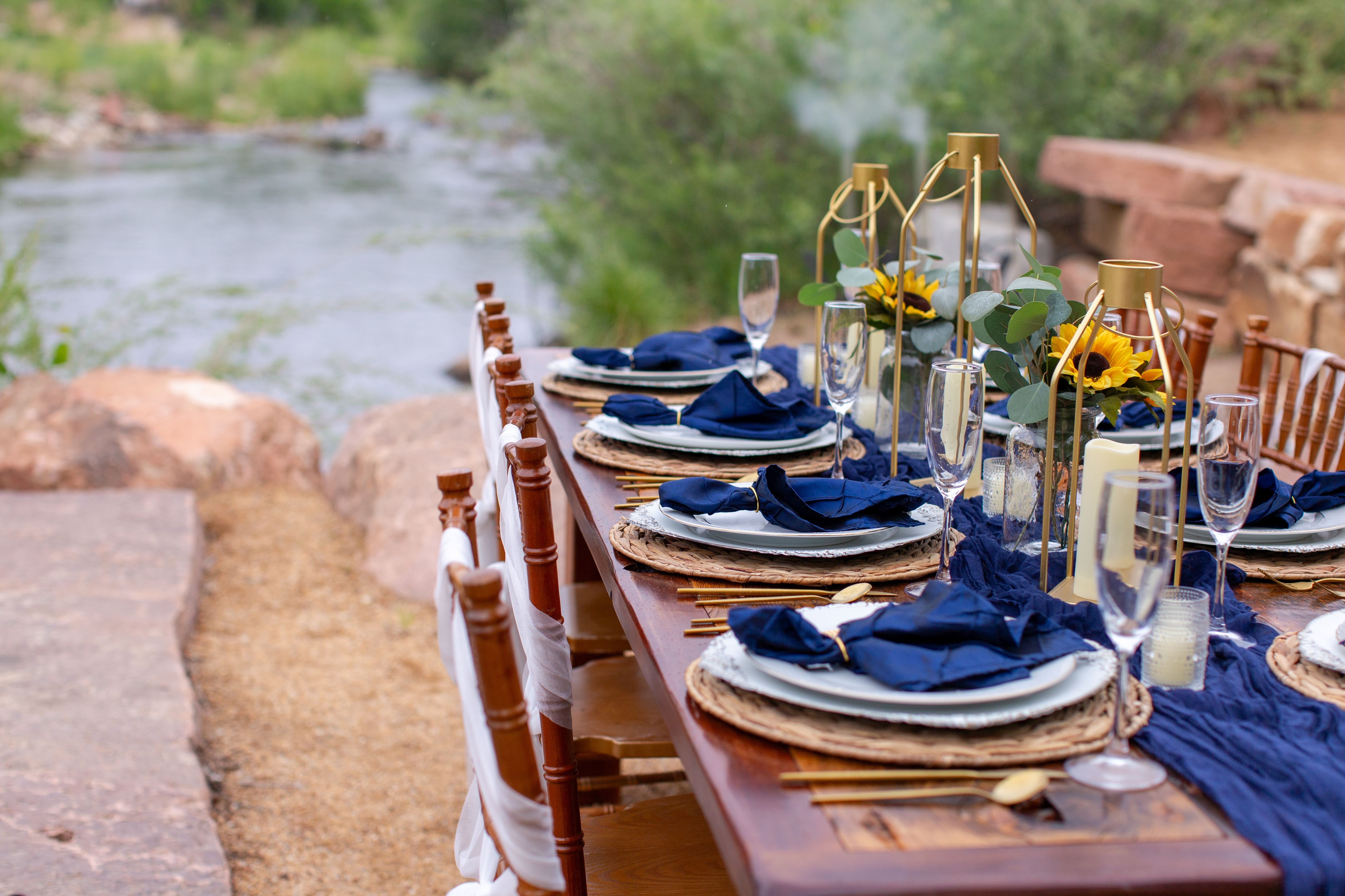 Outdoor dining table set with plates, glasses, and navy blue napkins near a river, with rocks and greenery in background.
