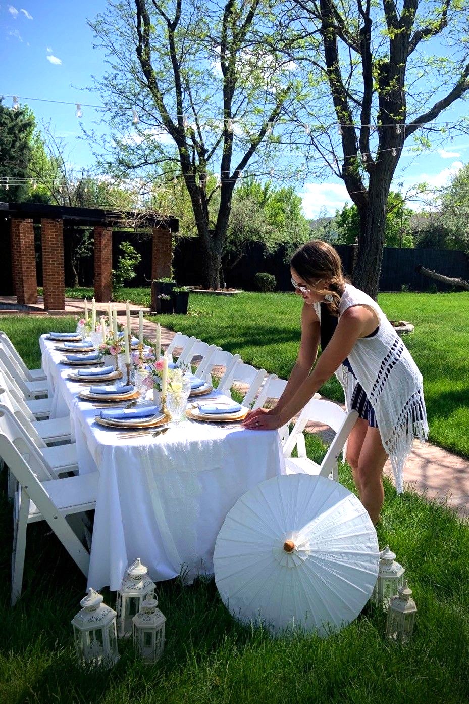 Woman arranging a table outdoors with white tablecloth, plates, and decorations, surrounded by trees and lanterns, sunny day.