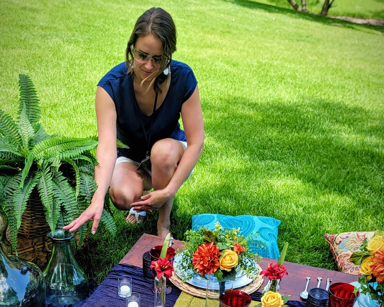 Woman crouching outdoors near a table with flowers and drinks, on a grassy lawn.