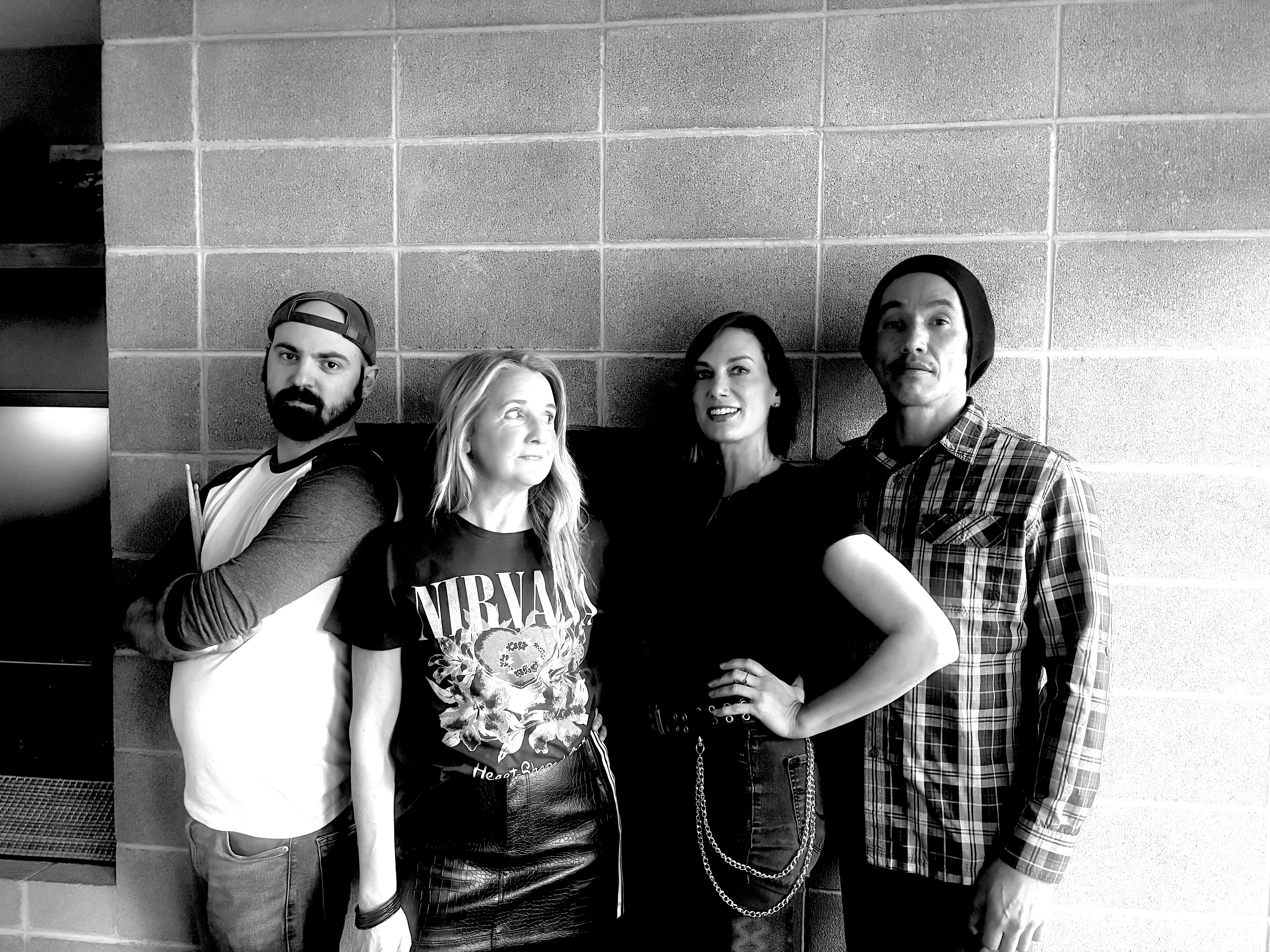 Four people standing against a tiled wall, posing for a photo, with varied clothing styles and expressions.