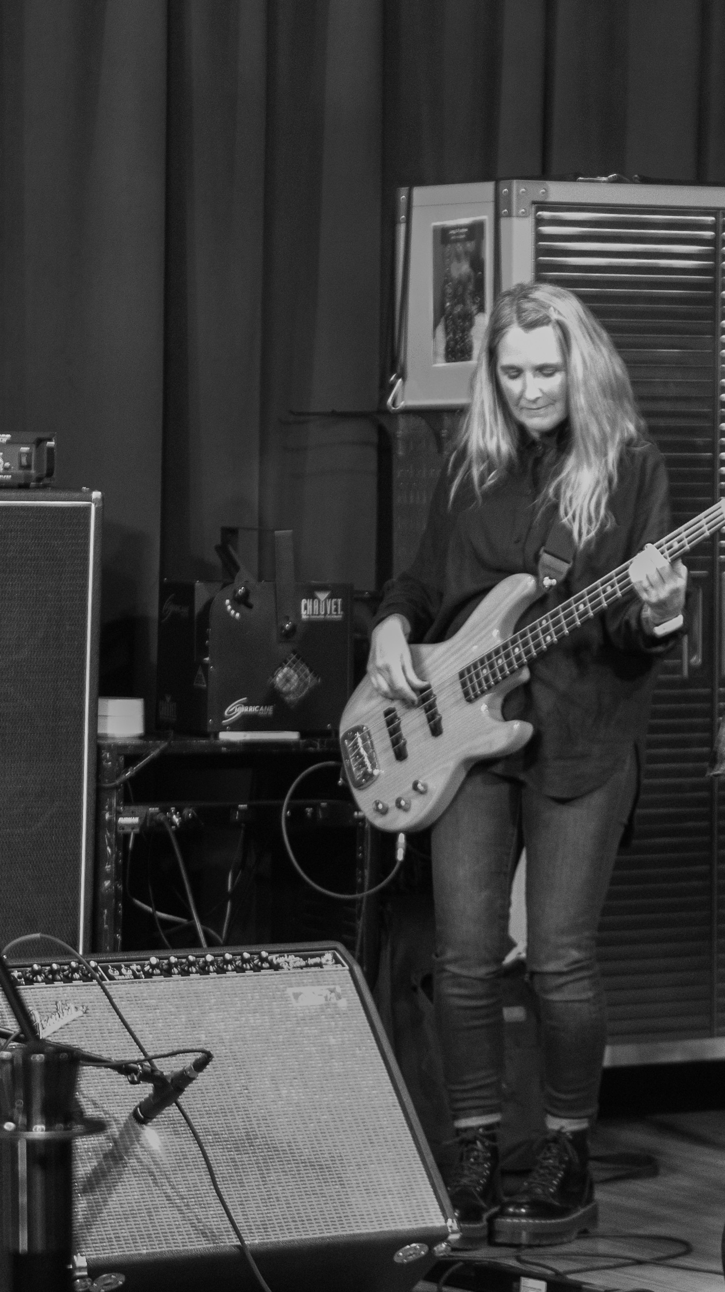 Woman with long hair playing bass guitar in a music studio, surrounded by equipment and a curtain backdrop.