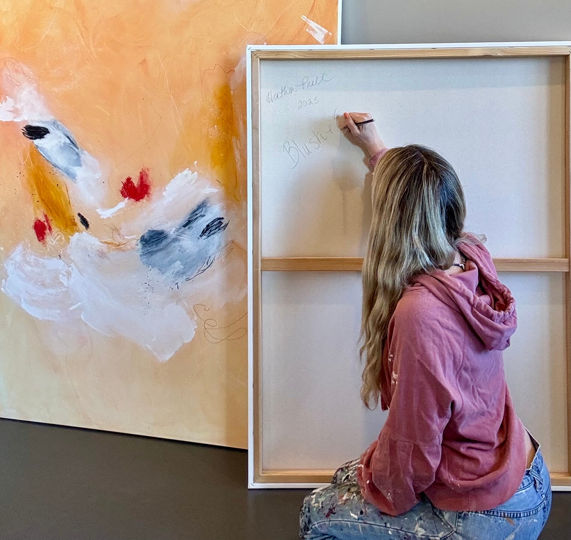 Girl with long hair in a pink hoodie sitting on the floor writing on a whiteboard, with a colorful abstract painting on the wall.
