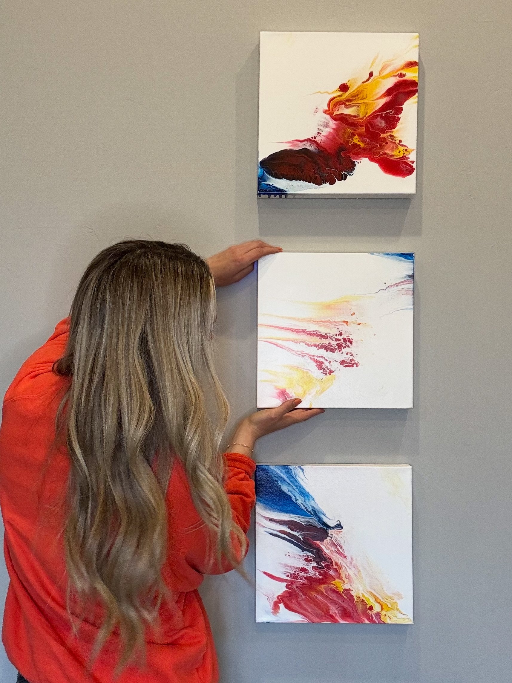 Person with long hair in red shirt reaching for three abstract paintings on a gray wall.