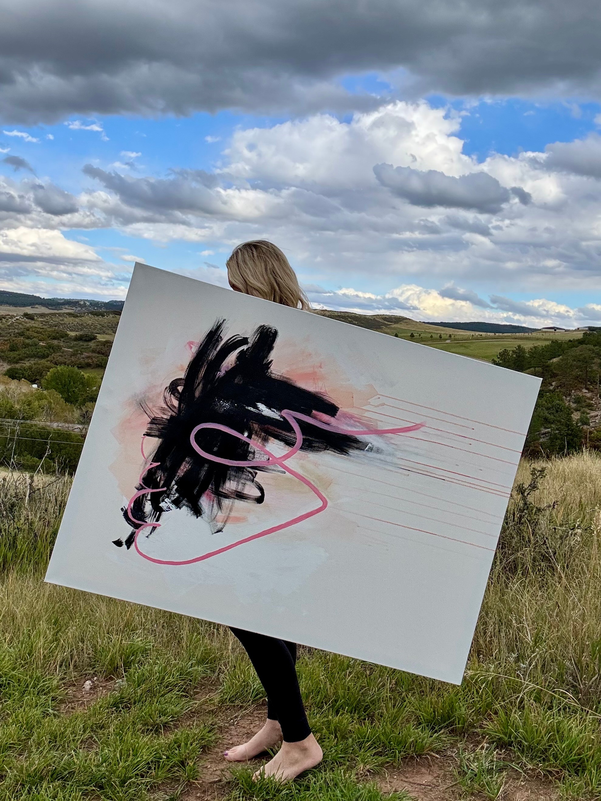 Person holding a large abstract painting outdoors with a cloudy sky and landscape background.