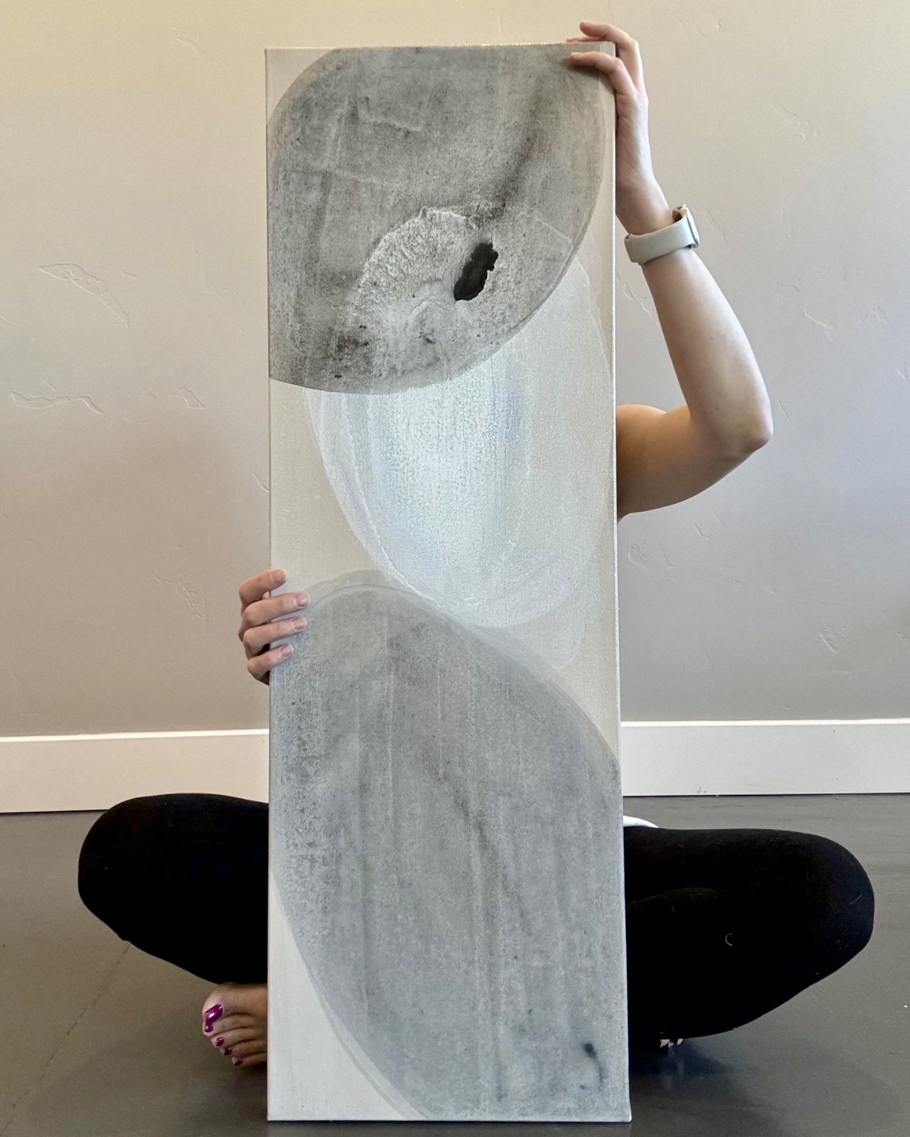 Person holding a tall abstract painting with gray, white, and black shapes, sitting on the floor against a plain wall.
