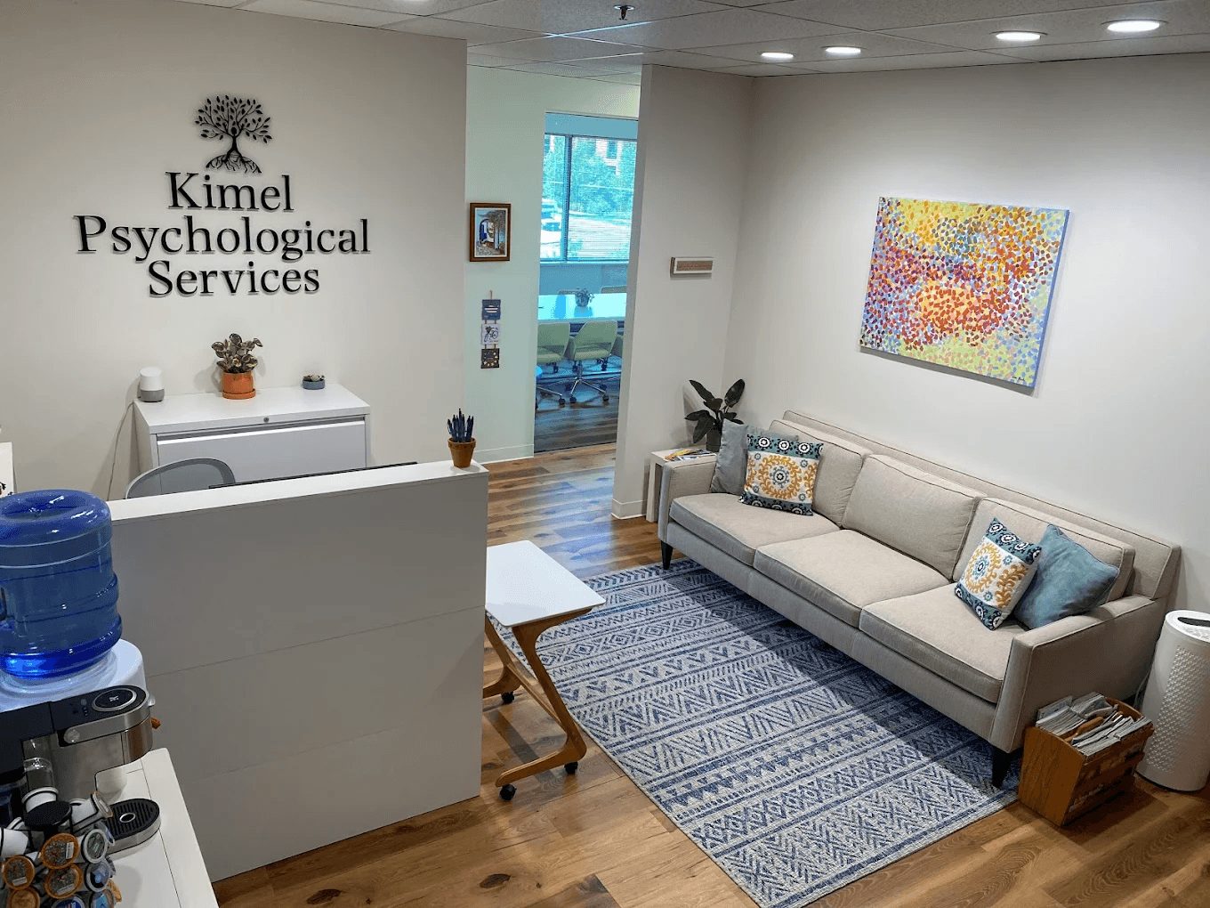Waiting room with a beige sofa, colorful artwork, and a patterned rug. A water dispenser and small table are visible. The wall has a sign reading 'Kimmel Psychological Services.'