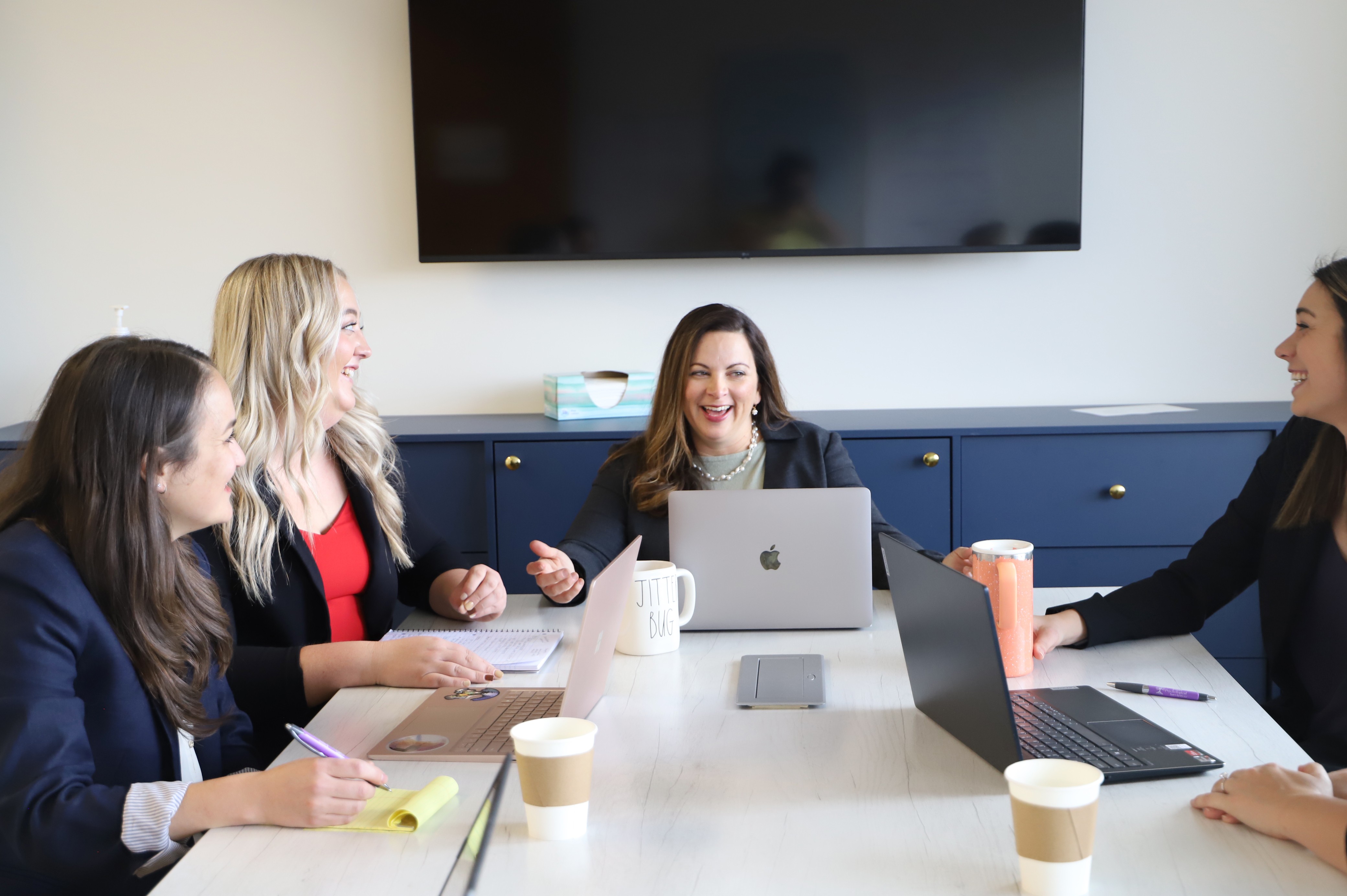 Five women sit around a conference table in a meeting room, smiling and engaging with laptops and notebooks.