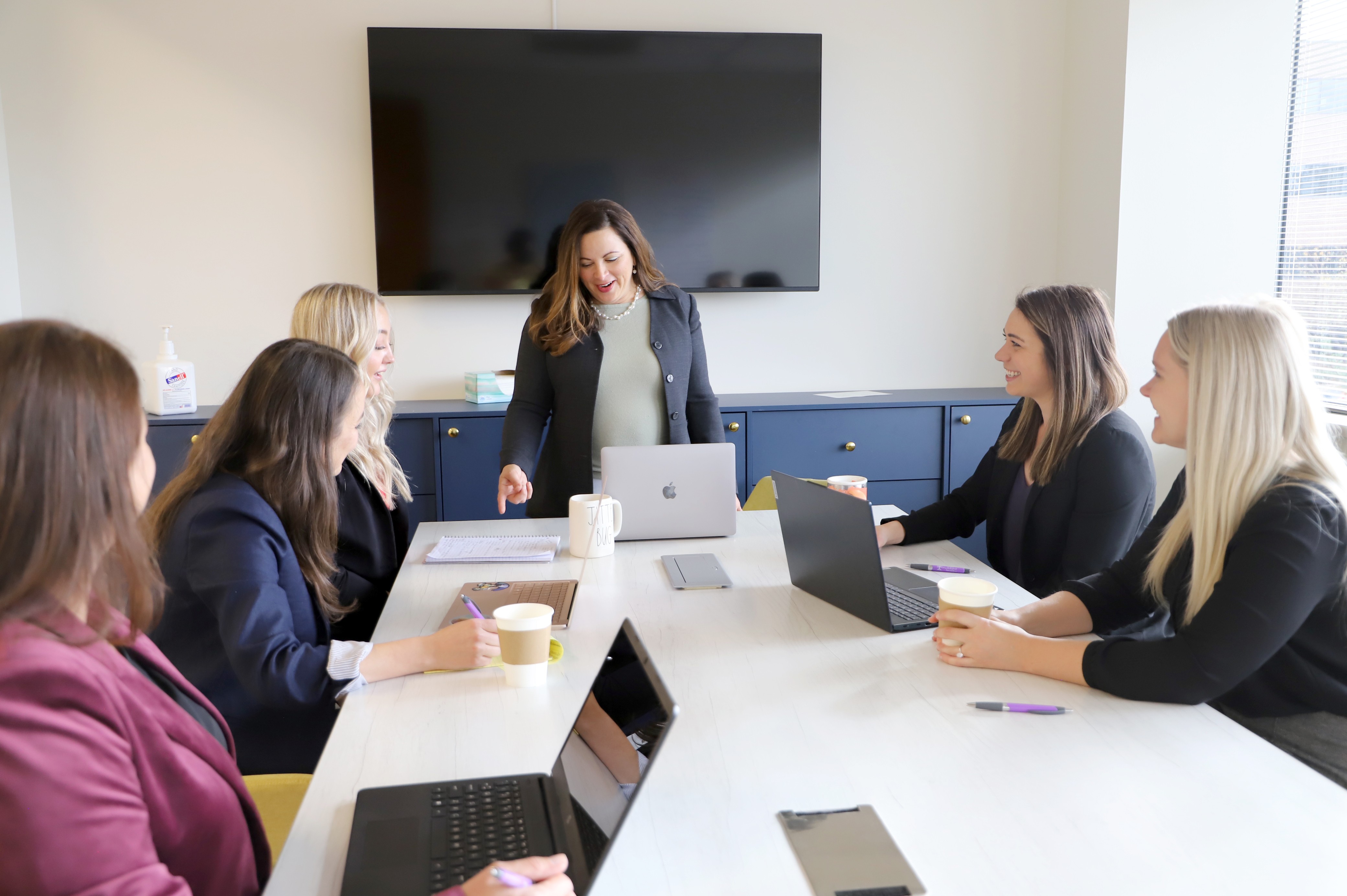 Six women sit around a conference table in a meeting room, with a woman standing at the head of the table.