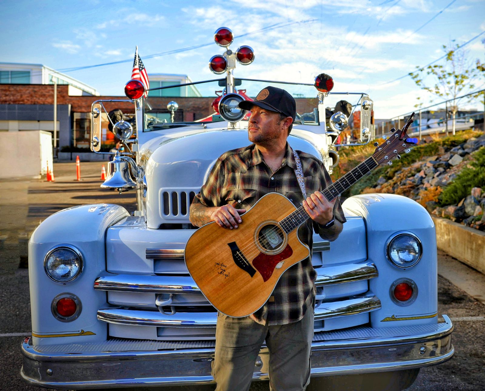 Man with a guitar standing in front of a vintage fire truck outdoors, with a blue sky and some trees.