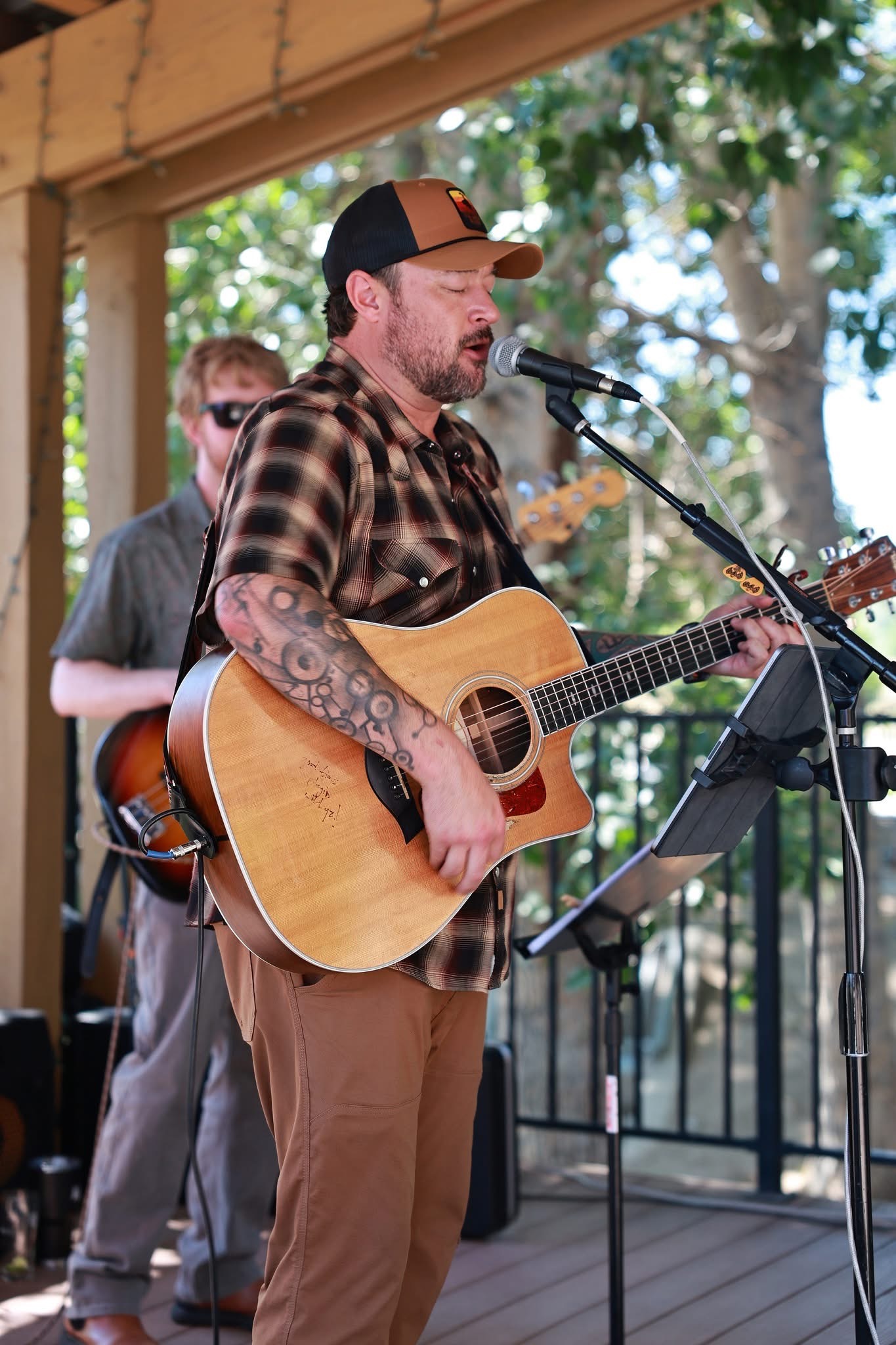 Man with tattoos playing acoustic guitar and singing into microphone outdoors, with trees in background.
