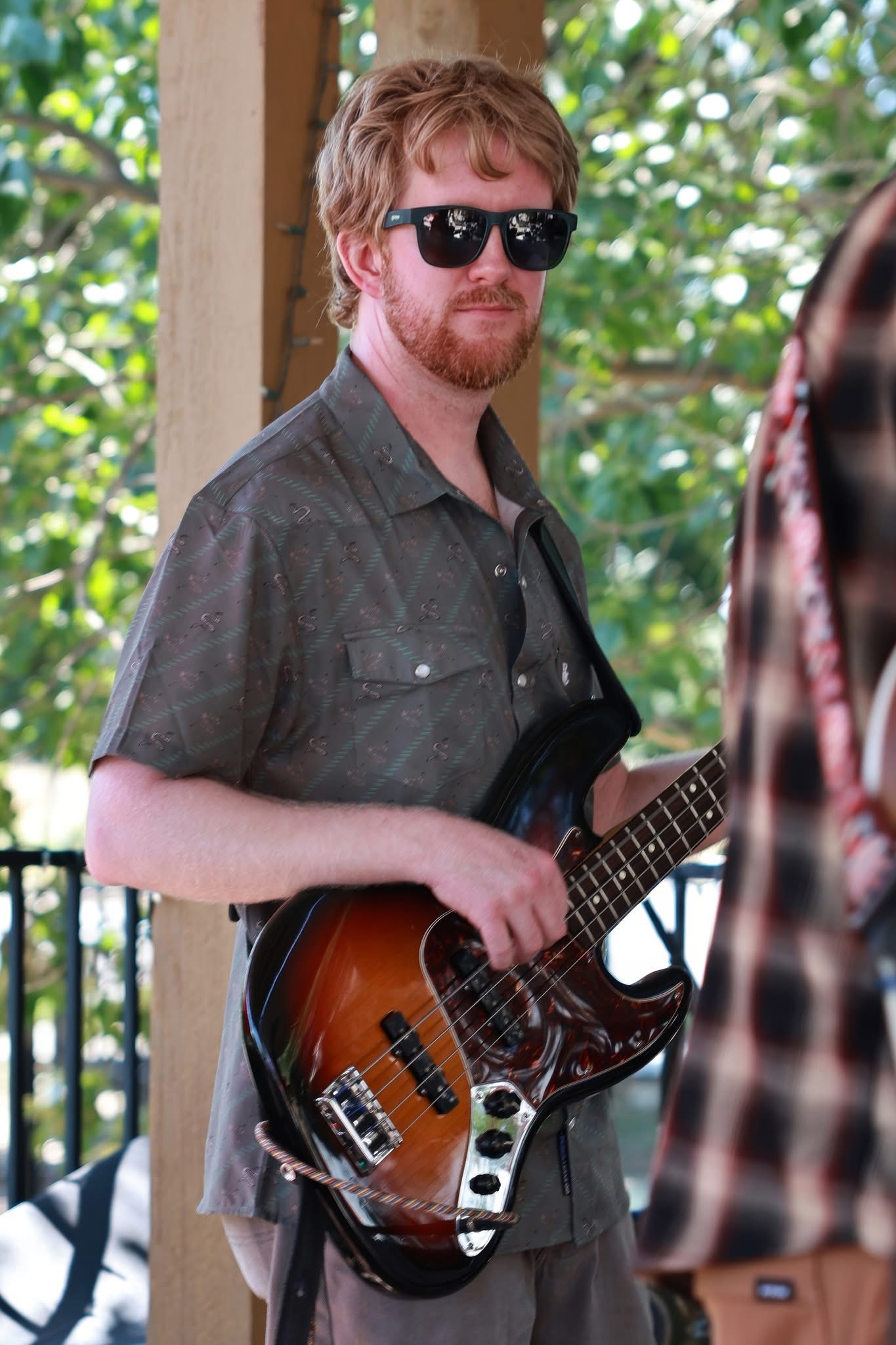 Young man with sunglasses holding an electric guitar outdoors, trees and a wooden structure in background.
