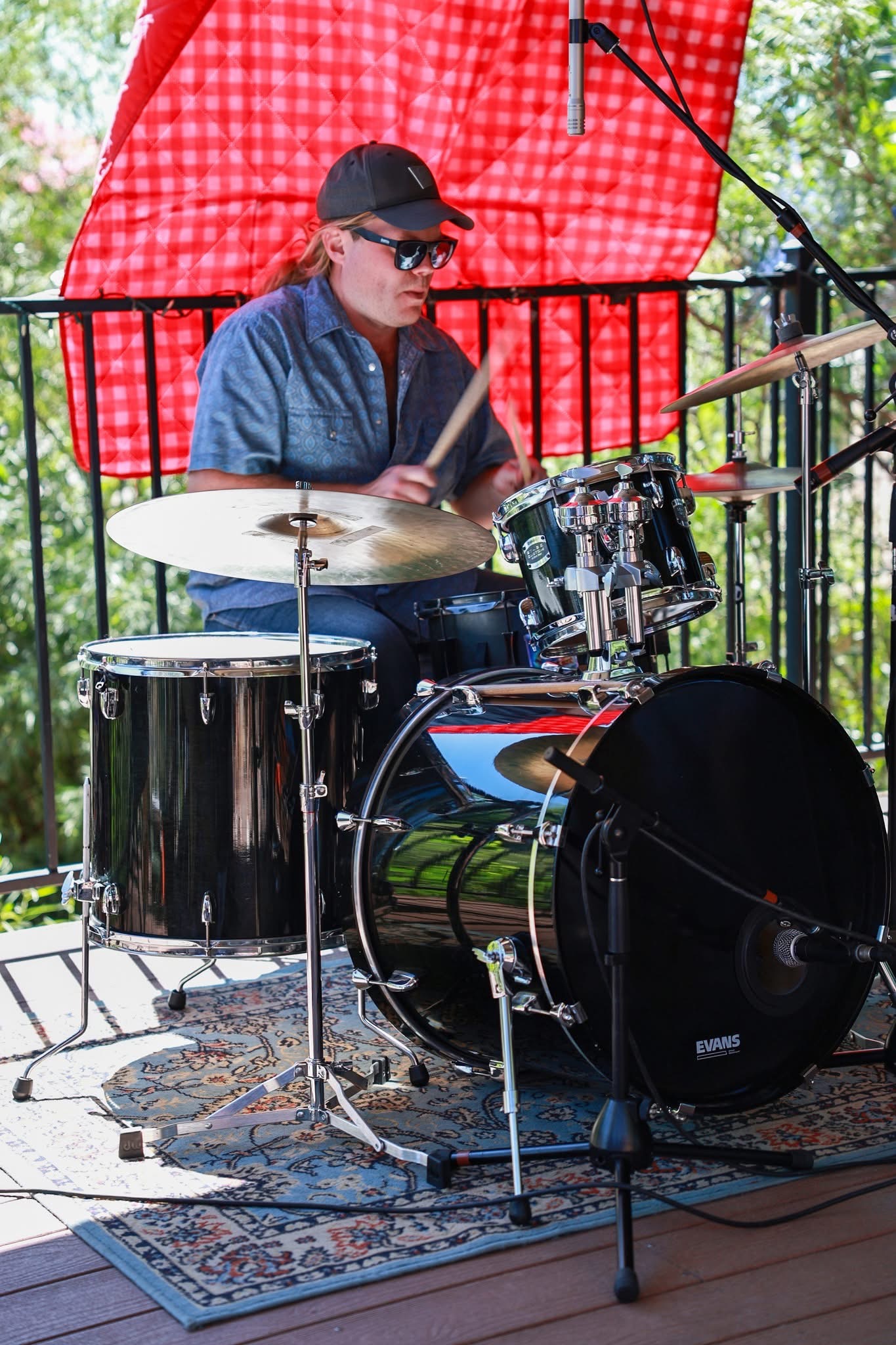 Person playing drums outdoors on a stage with a red umbrella and trees in background.