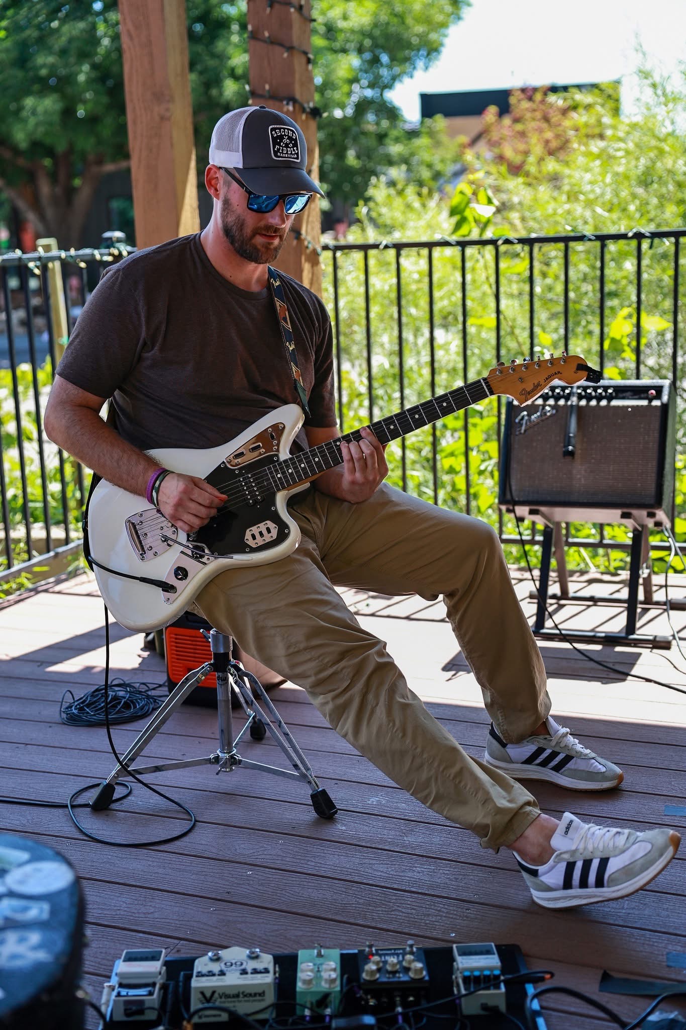 Man playing electric guitar outdoors on a wooden deck with a small amplifier and pedalboard, wearing sunglasses, a cap, and casual clothing.