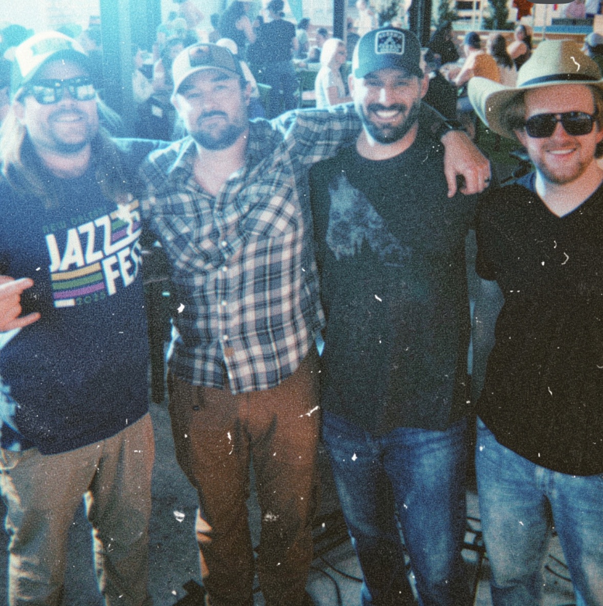 Four men standing together outdoors, smiling, with arms around each other, wearing casual clothing and hats.