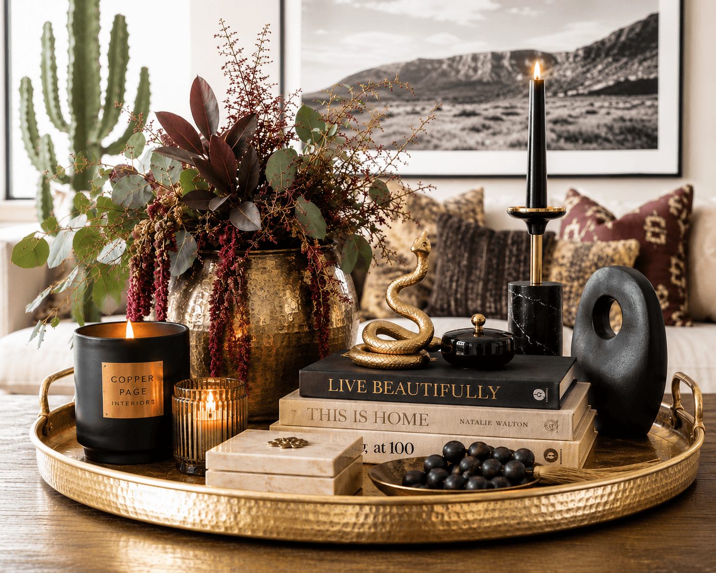 Decorative tray with candles, a vase with flowers, a black sculpture, books, and a bowl of black berries on a table.