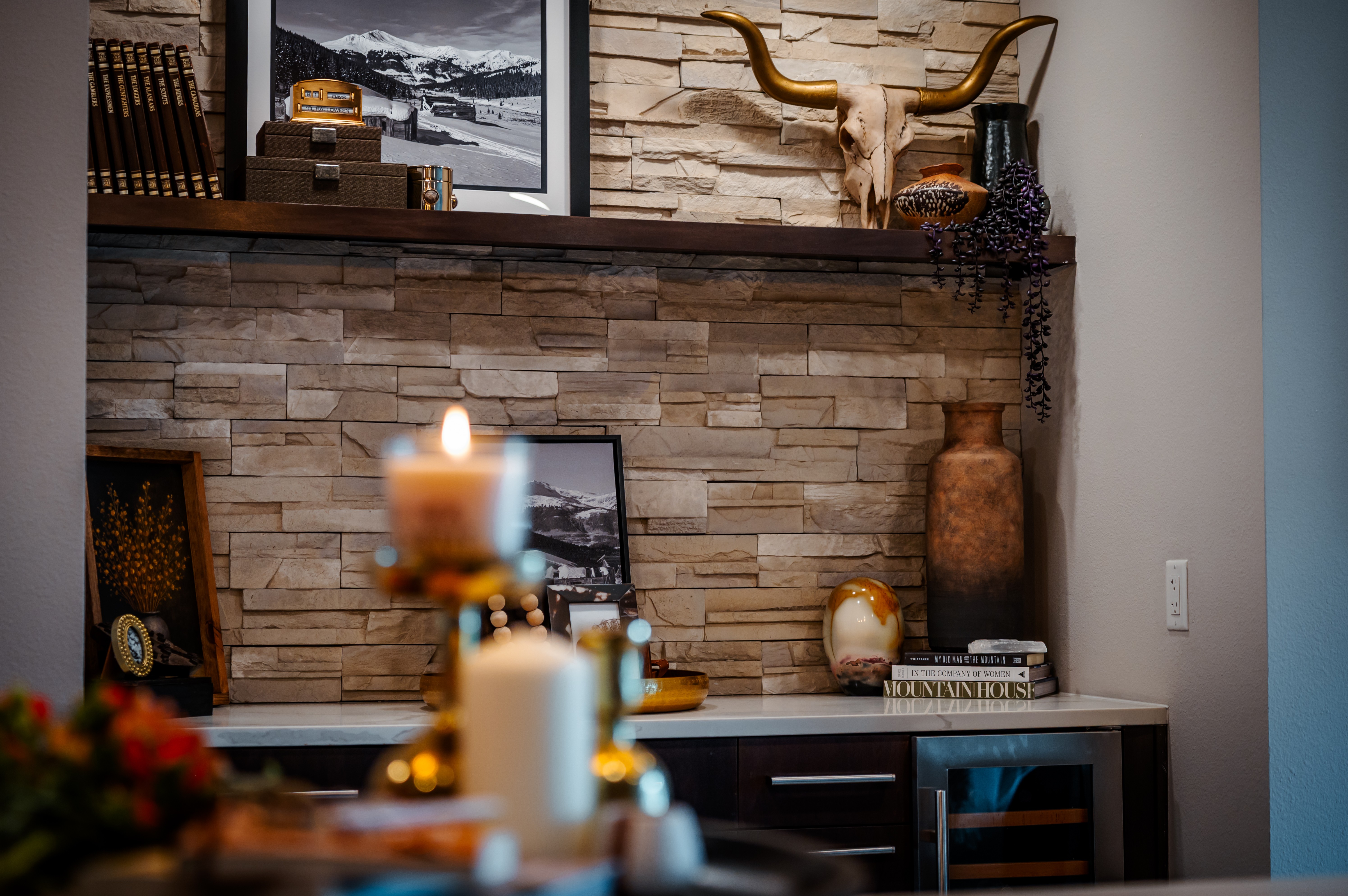 Decorative interior with candles, framed pictures, and a stone wall, featuring a large vase and a shelf with various objects.