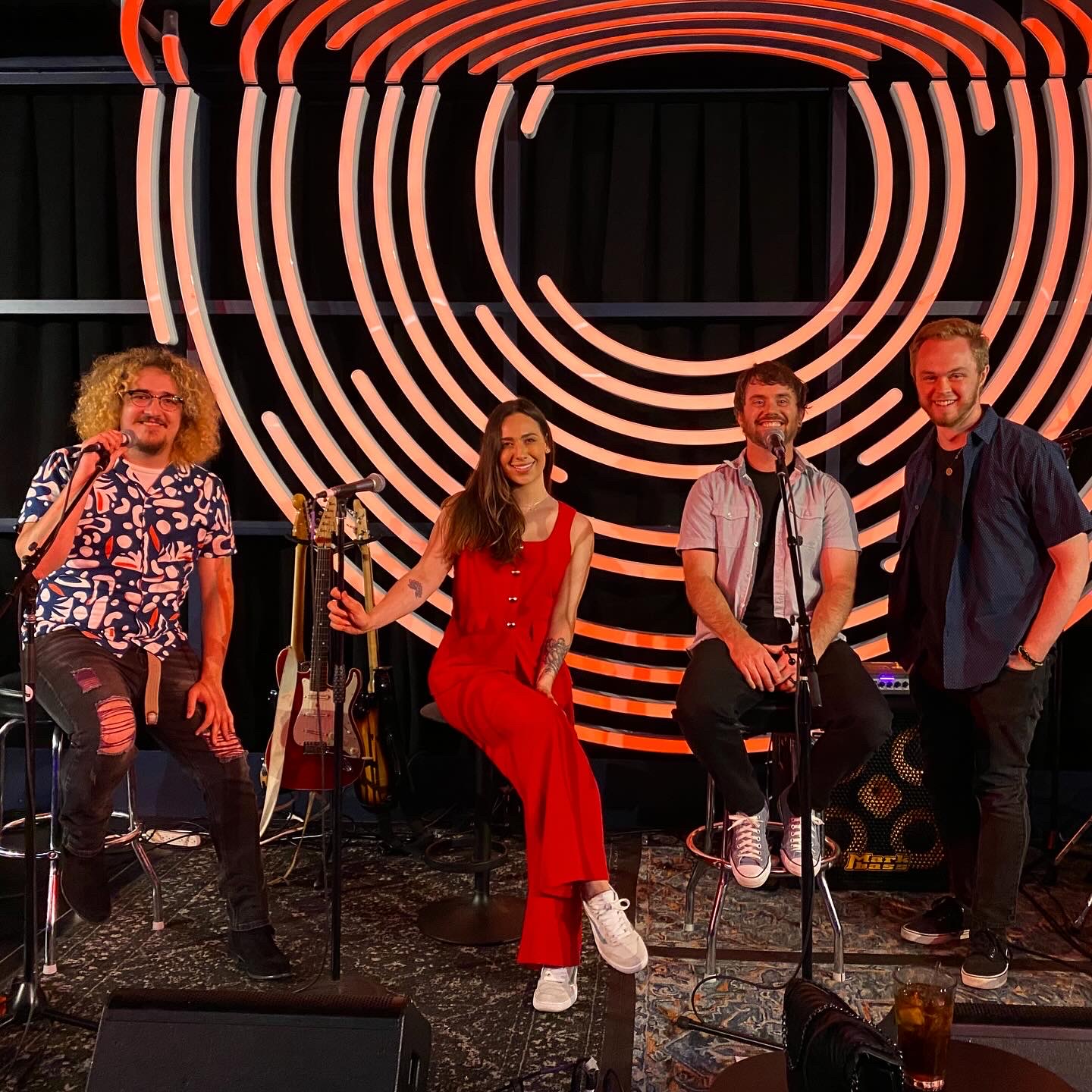 Four people on a stage with a circular neon light backdrop, some holding musical instruments, smiling at the camera.