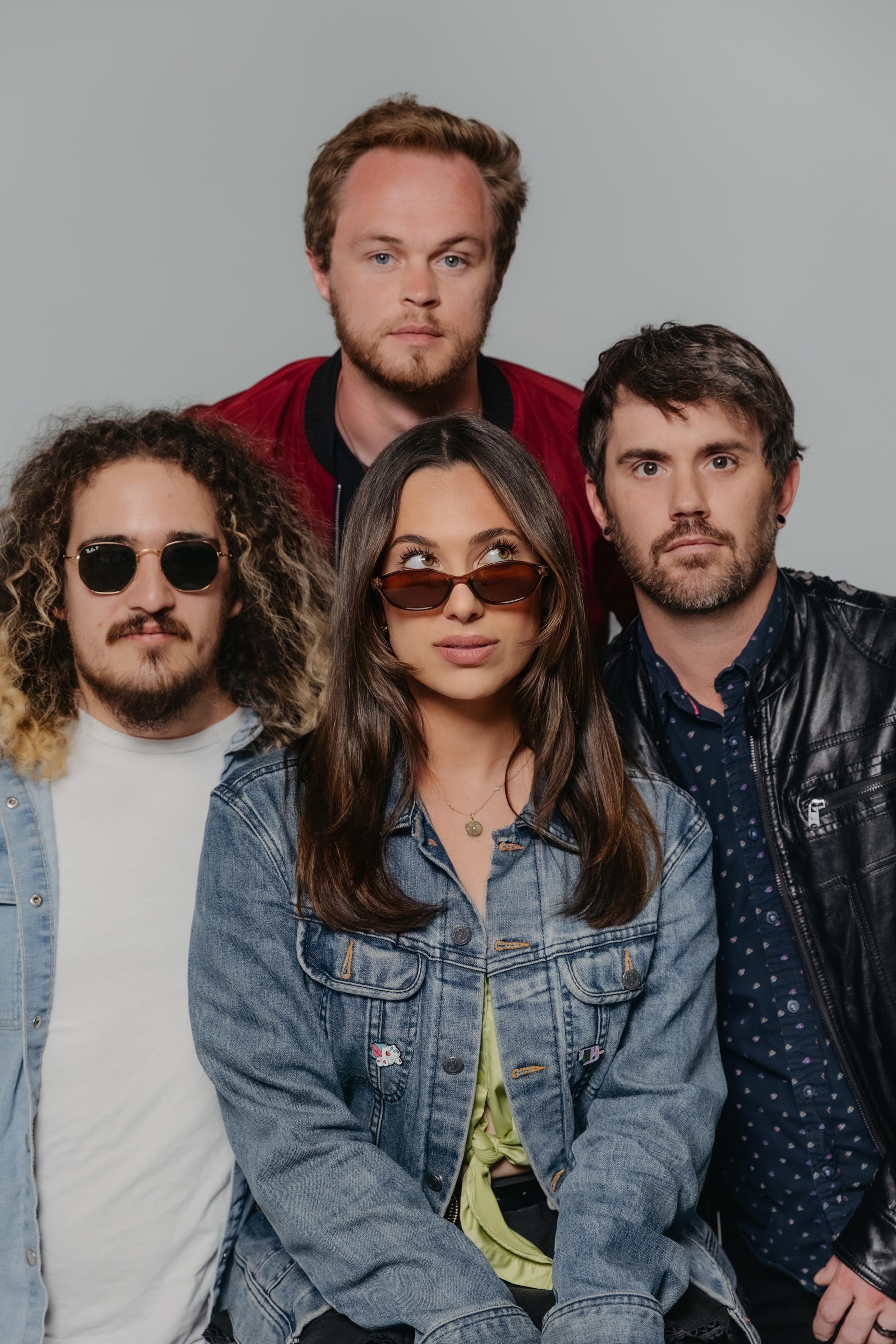 Four people pose together, two women and two men, against a plain background, wearing casual clothing and sunglasses.