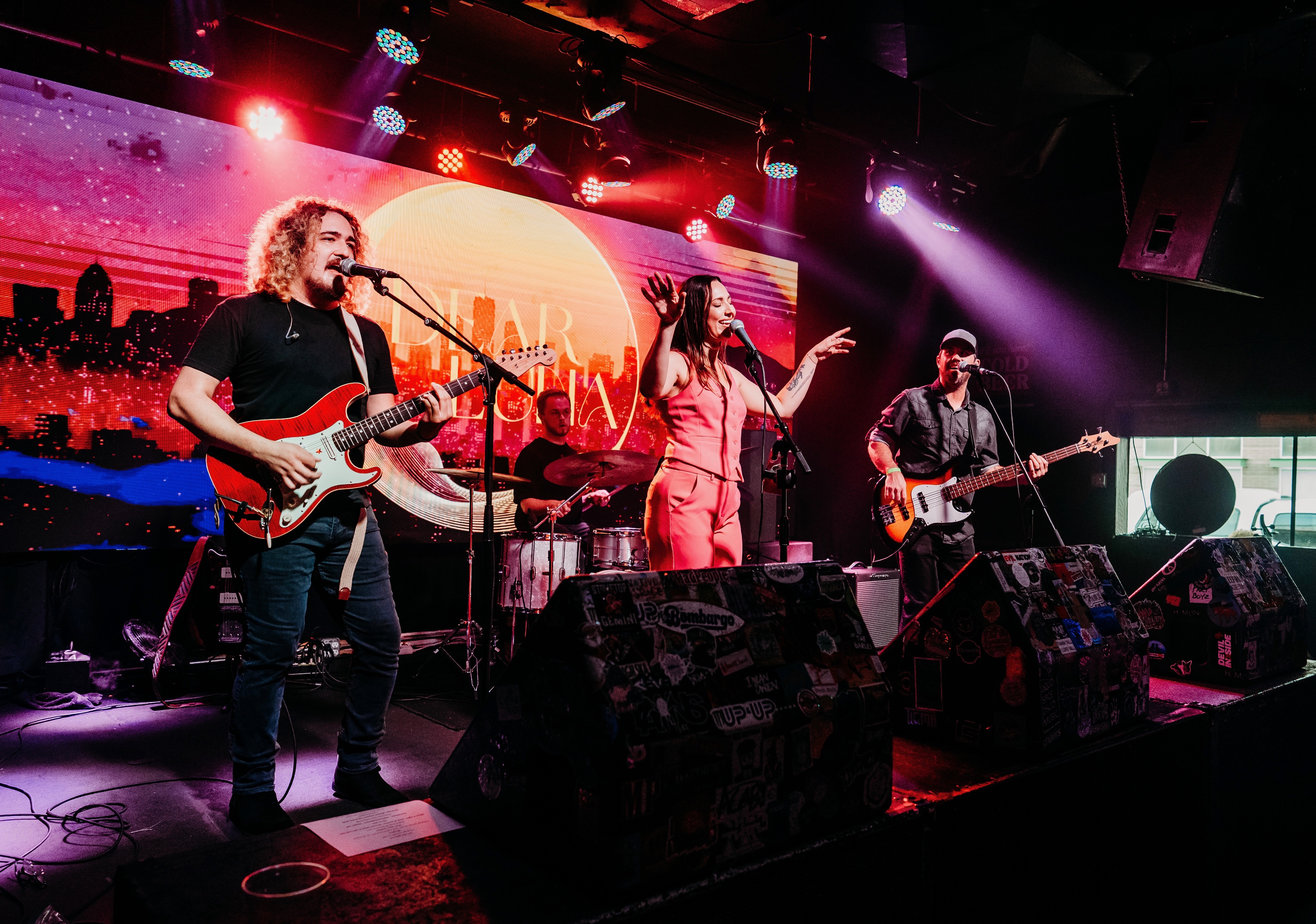 Three musicians perform on stage with a colorful cityscape background, playing guitars and singing, under stage lights.