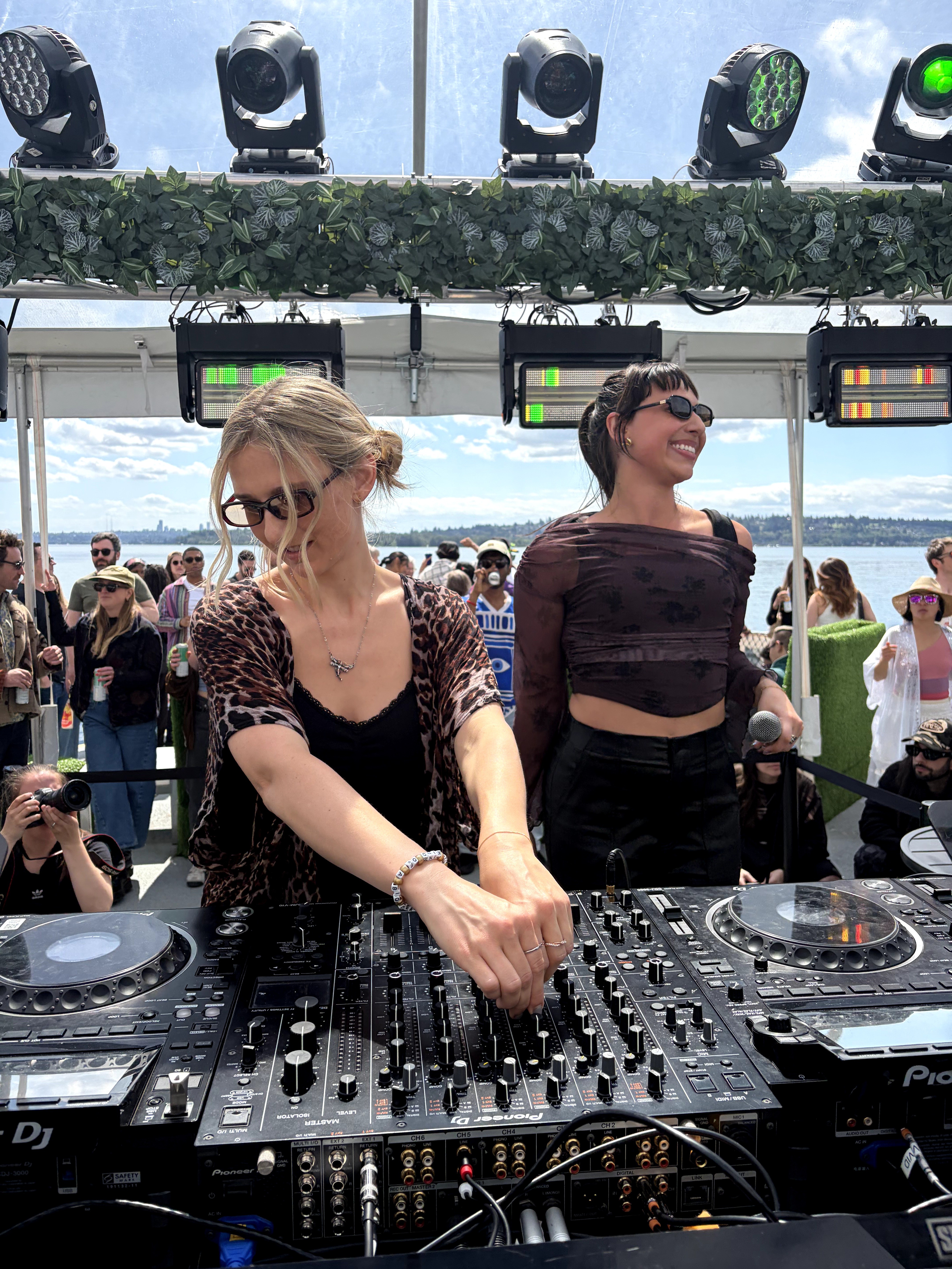 Two women DJing outdoors with a crowd and sky in the background, one adjusting equipment, the other smiling, wearing sunglasses.
