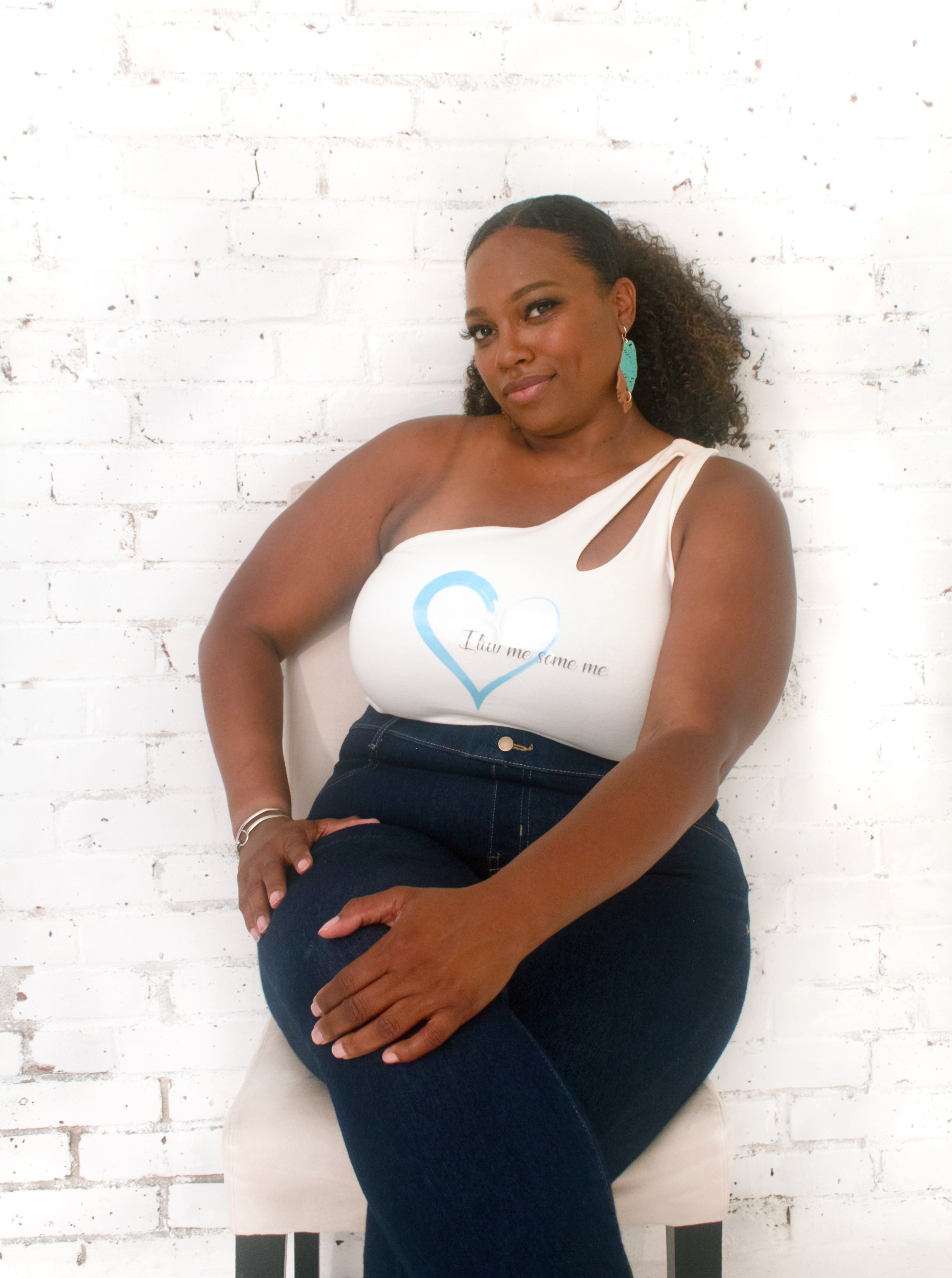 Woman sitting on a chair against a white brick wall, wearing a white top and dark pants, smiling.