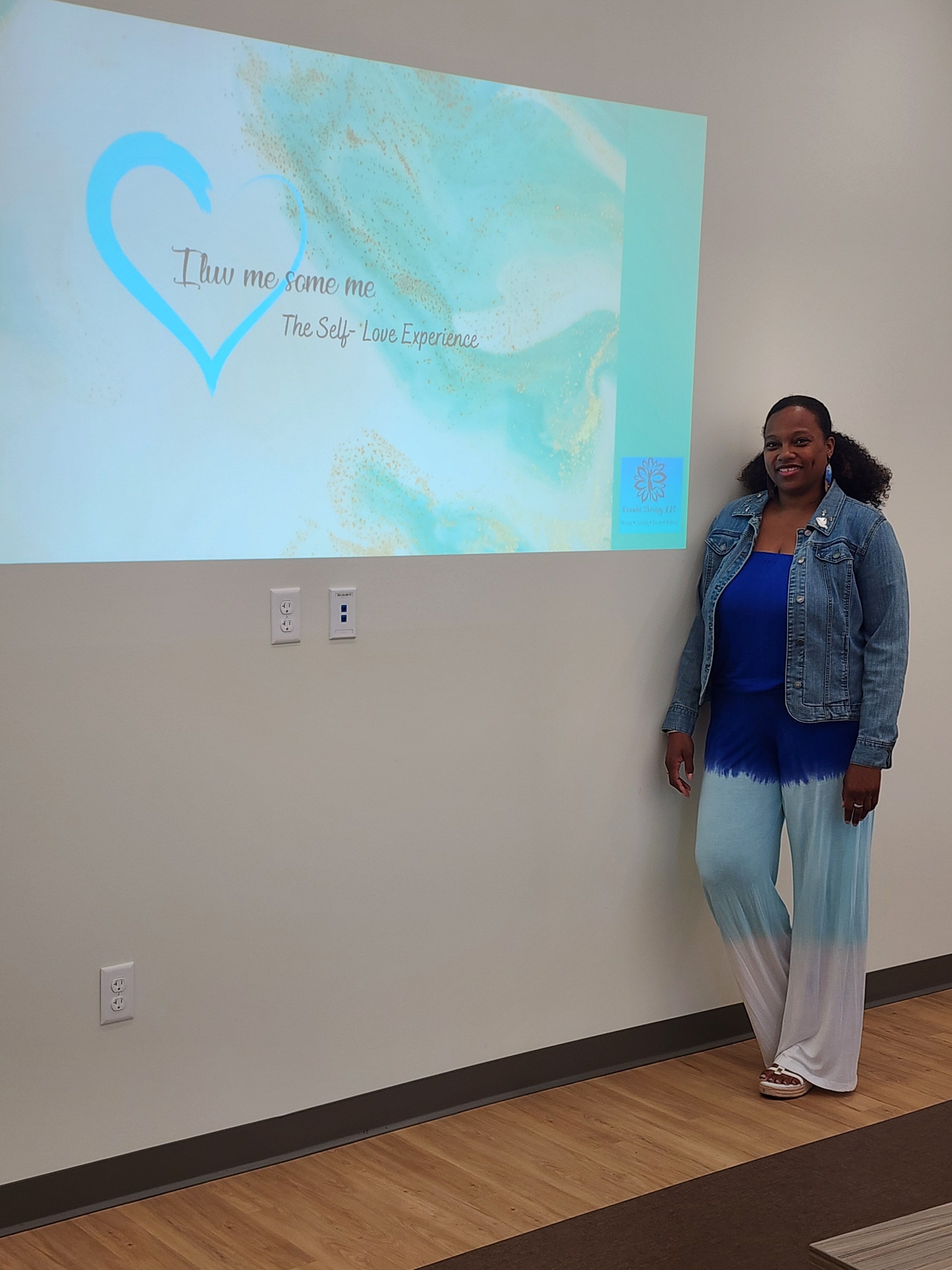 Person standing next to a wall with a projected image of a blue and green abstract design, wearing a denim jacket and white pants.